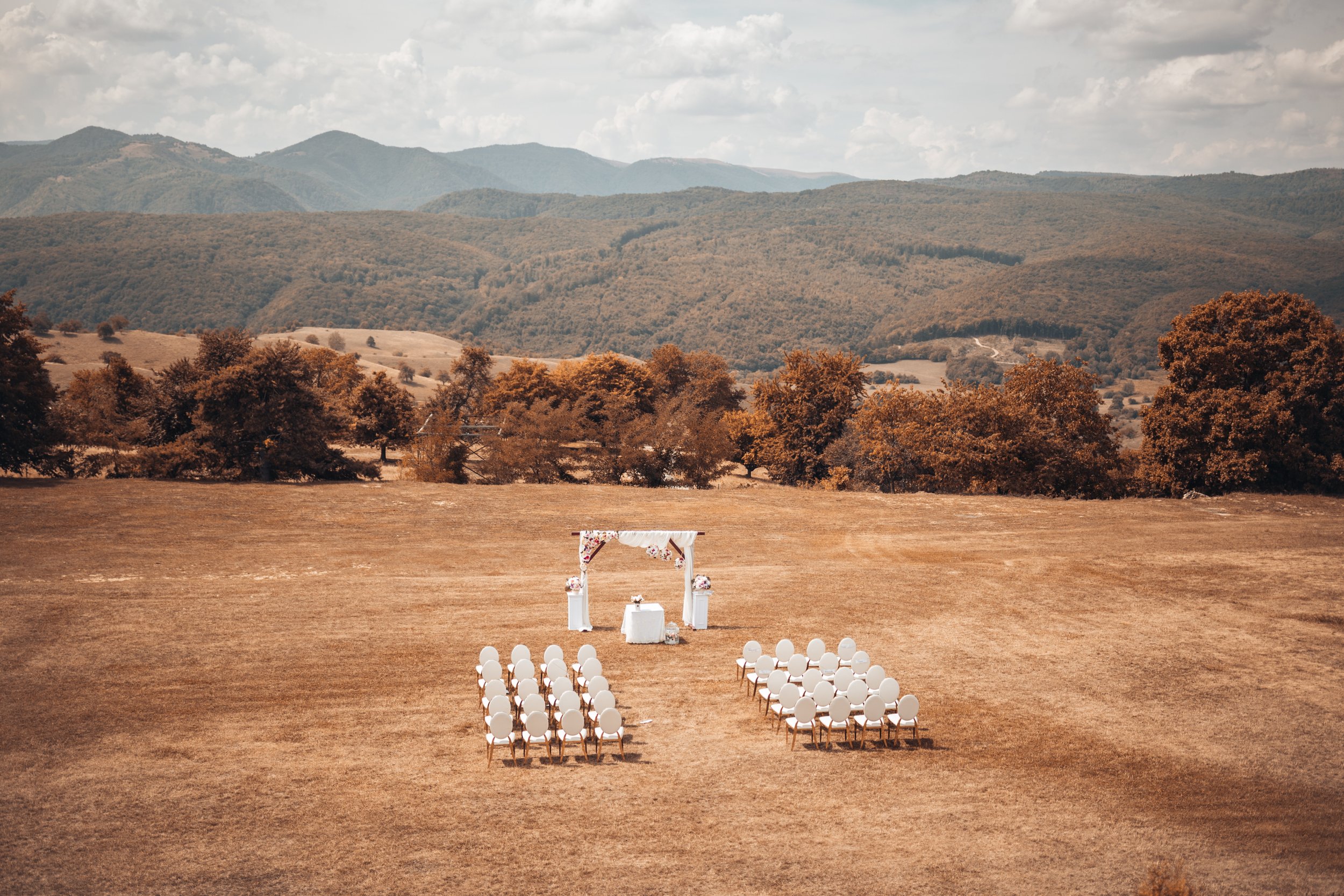 Outdoor wedding ceremony setup on a dry grassy field with white chairs arranged in rows facing a decorated archway, with mountains and a cloudy sky in the background.