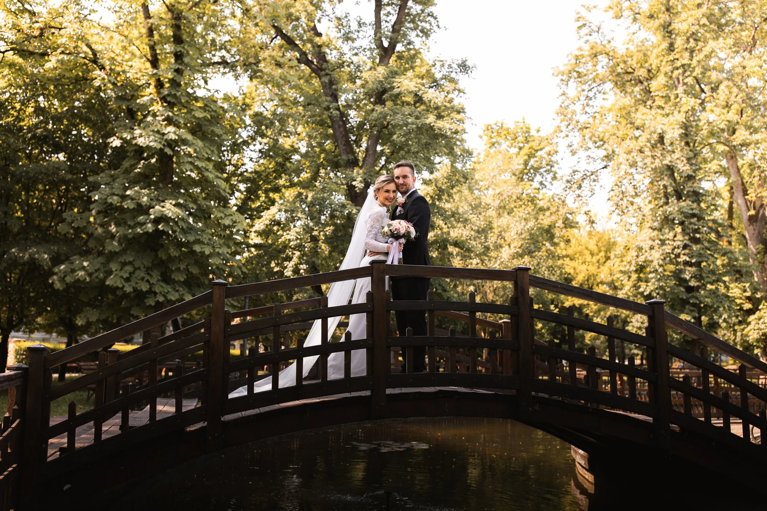 A bride and groom standing on a small wooden bridge over water in a park with green trees and sunlight in the background.