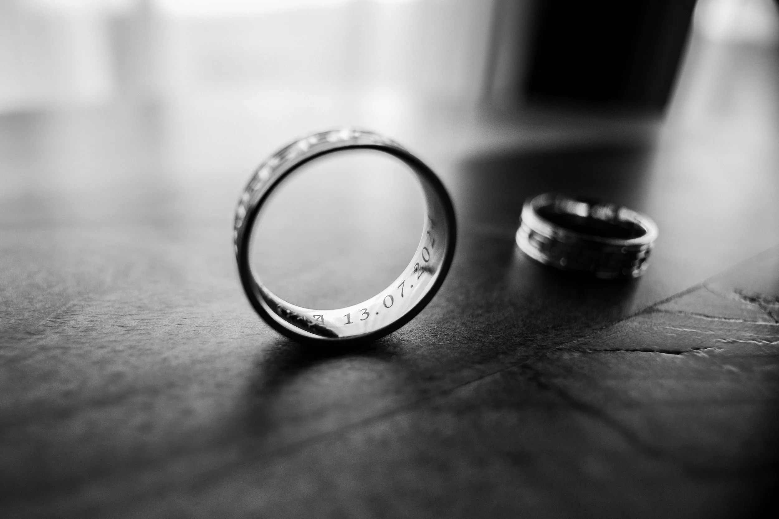 Close-up shot of a metallic ring with engraved markings, lying on a textured surface with another ring blurred in the background, in black and white.