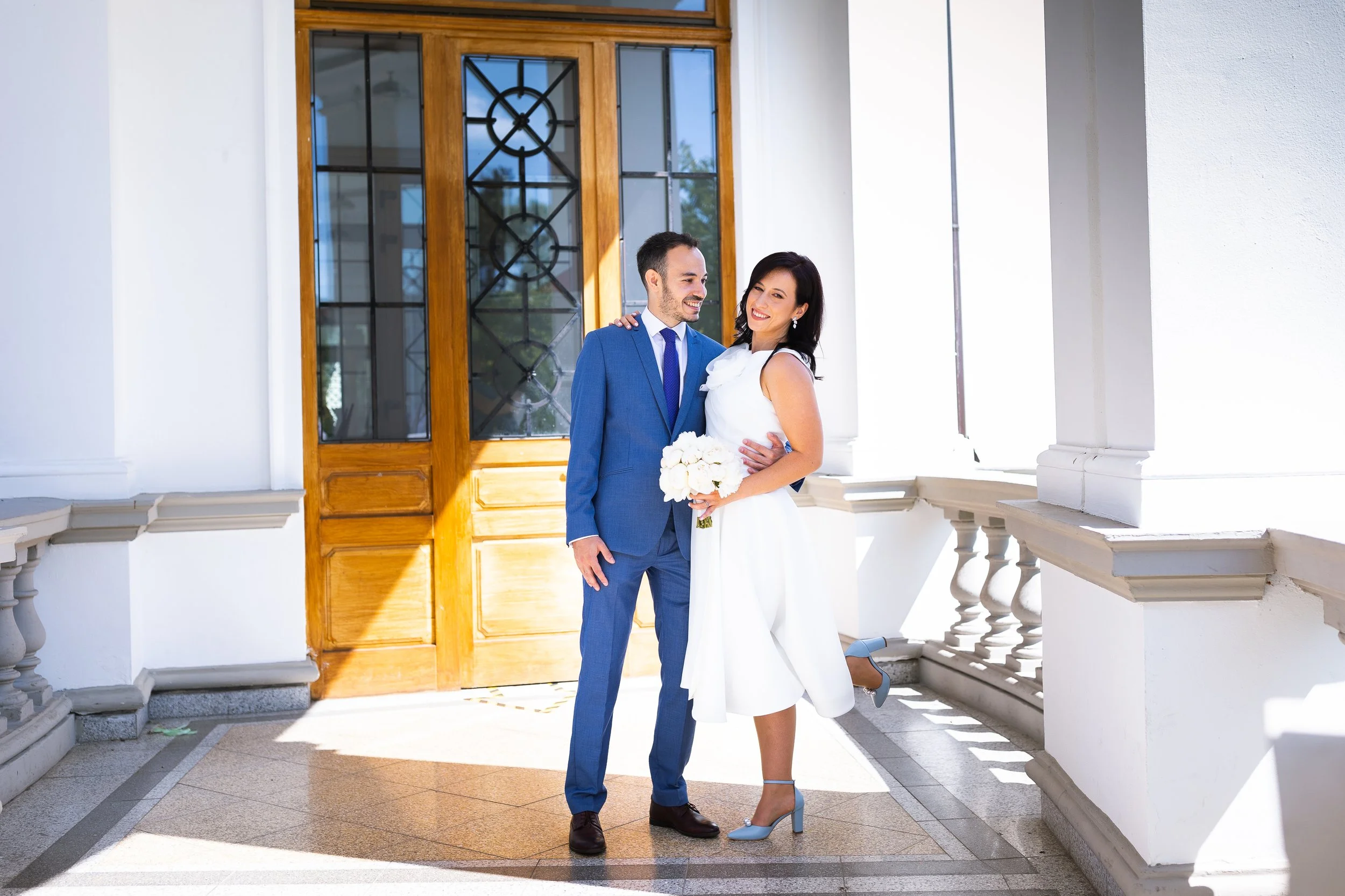A smiling couple dressed in wedding attire, the bride in a white dress holding a bouquet and the groom in a blue suit, standing on a porch with white columns and a wooden door with glass panels.
