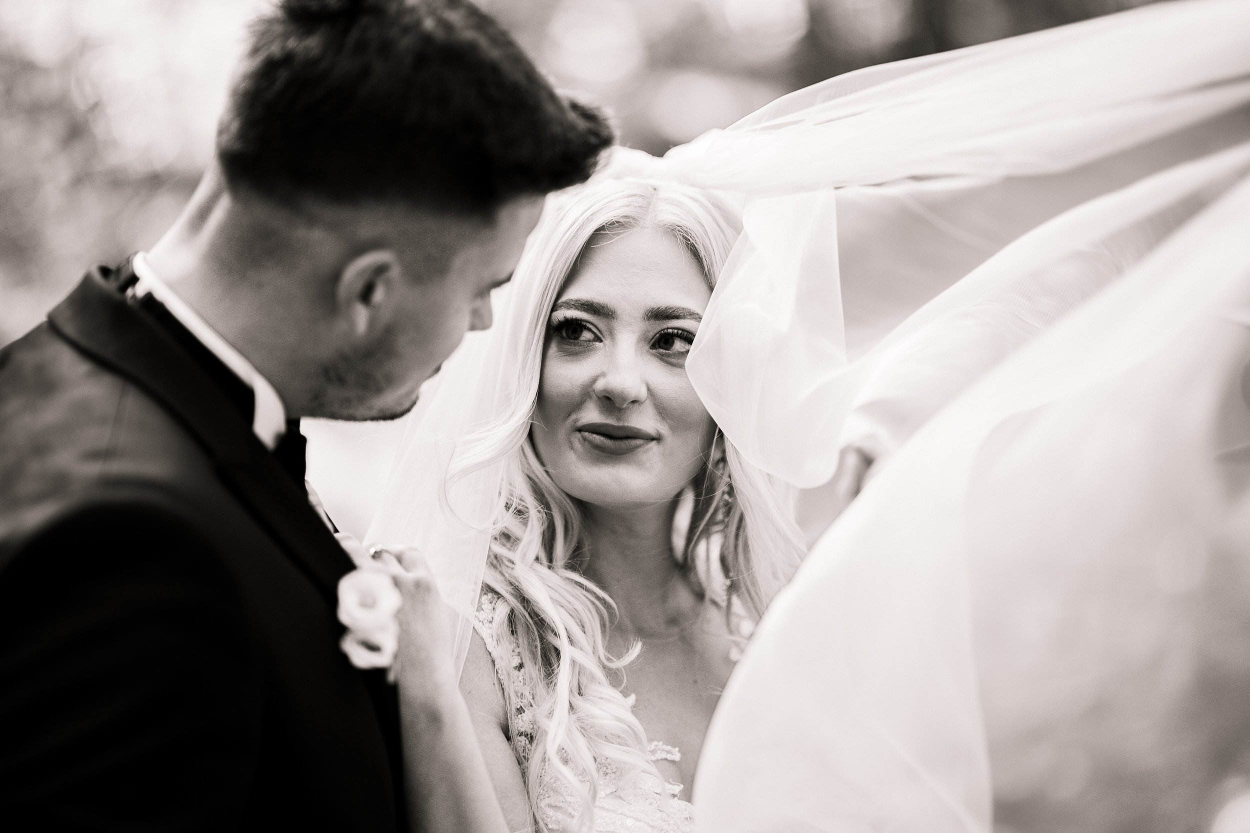 A bride and groom having a close moment outdoors, with the bride looking at the groom and the groom looking down, both dressed in wedding attire.