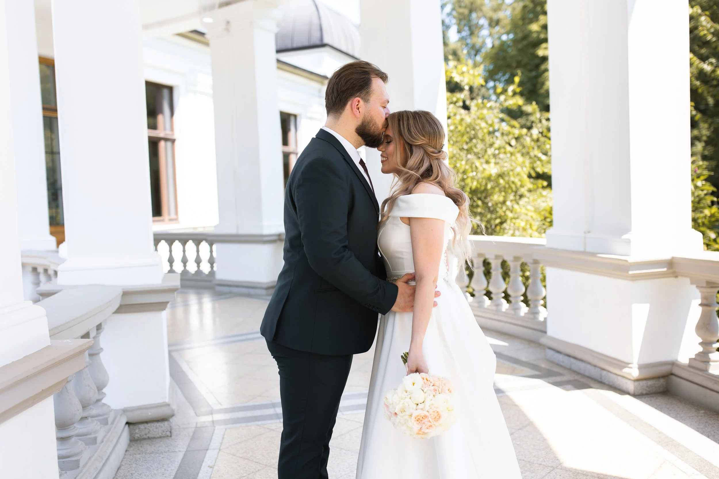 A bride and groom stand close together on a white porch with columns, sharing a tender moment. The bride is in a white wedding dress holding a bouquet of flowers, and the groom in a black suit and tie.