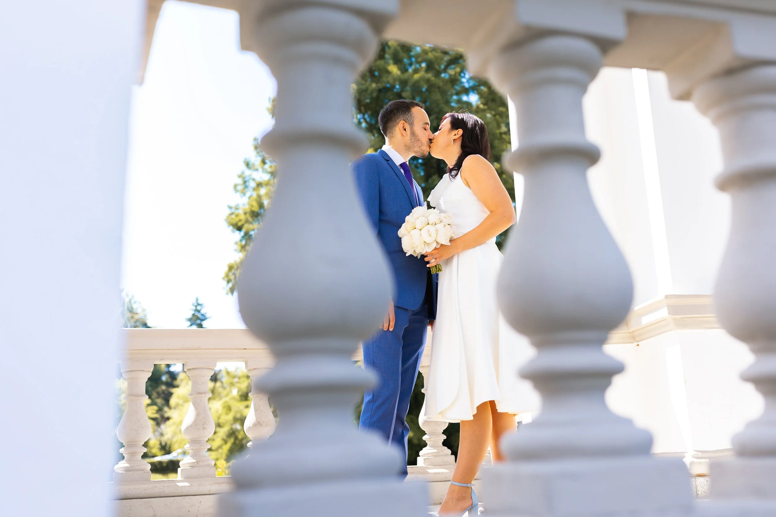 A bride and groom kissing during their wedding, viewed through white balcony balusters, with trees and a blue sky in the background.