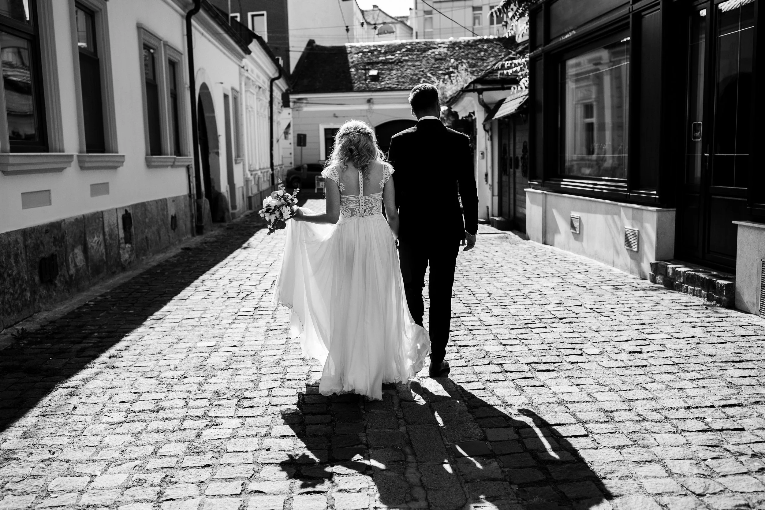 A black and white photo of a bride and groom walking down a cobblestone street, holding hands, with the bride holding a bouquet of flowers.