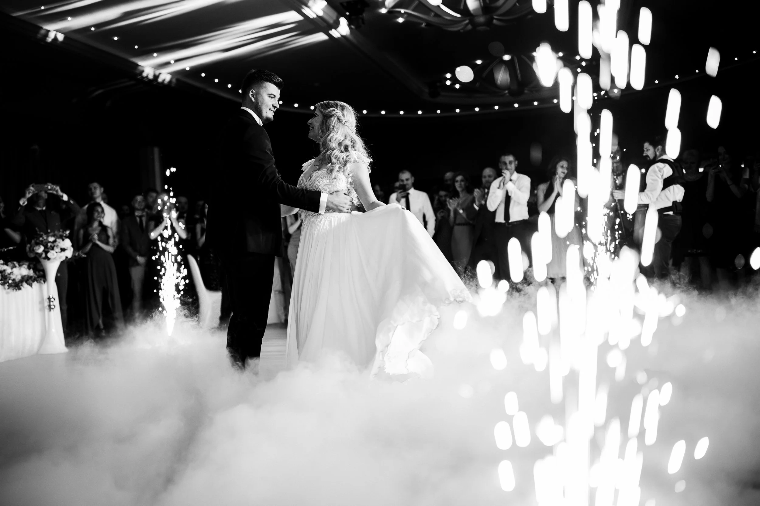 Black and white photo of a bride and groom dancing during their wedding reception, surrounded by guests. The bride is wearing a long wedding gown, and the groom is in a suit. There are sparklers and fog on the dance floor with decorative lights overh