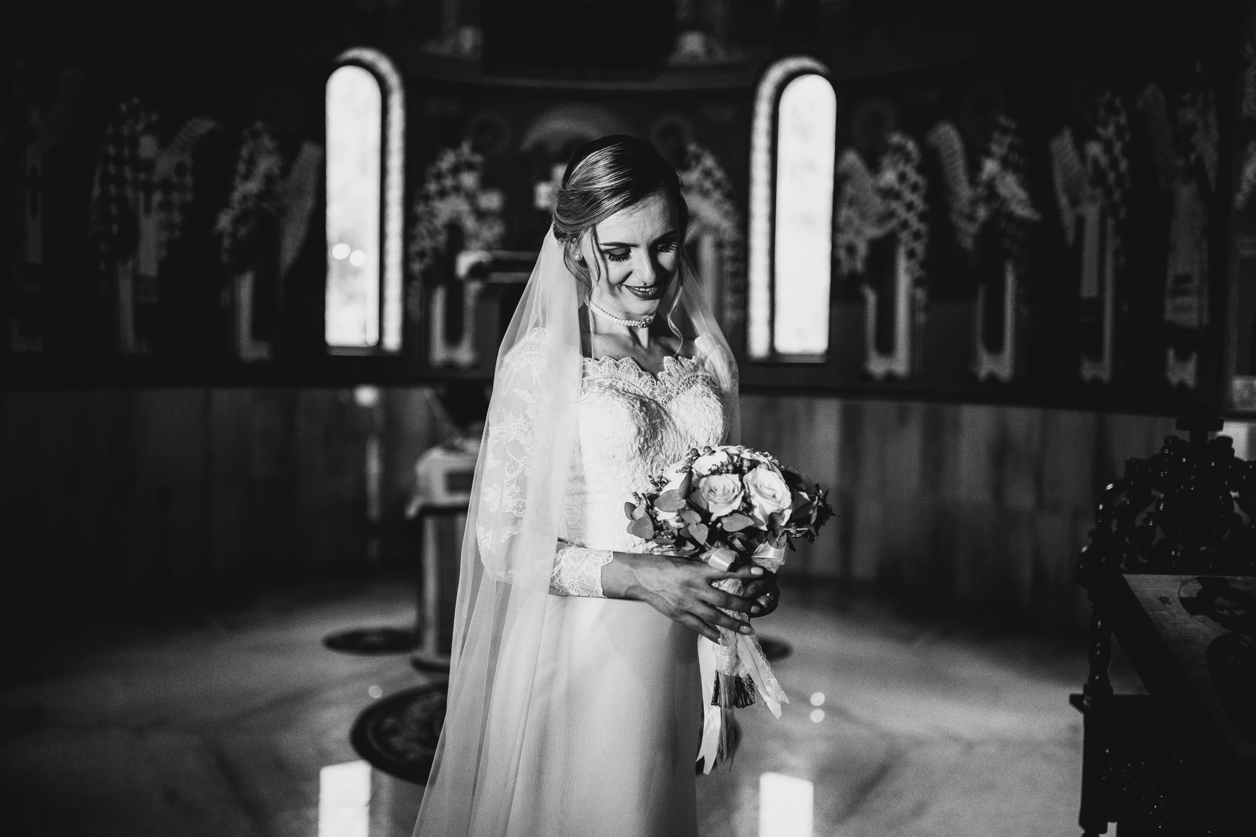 A bride in a lace wedding dress holding a bouquet of roses inside a church with stained glass windows.