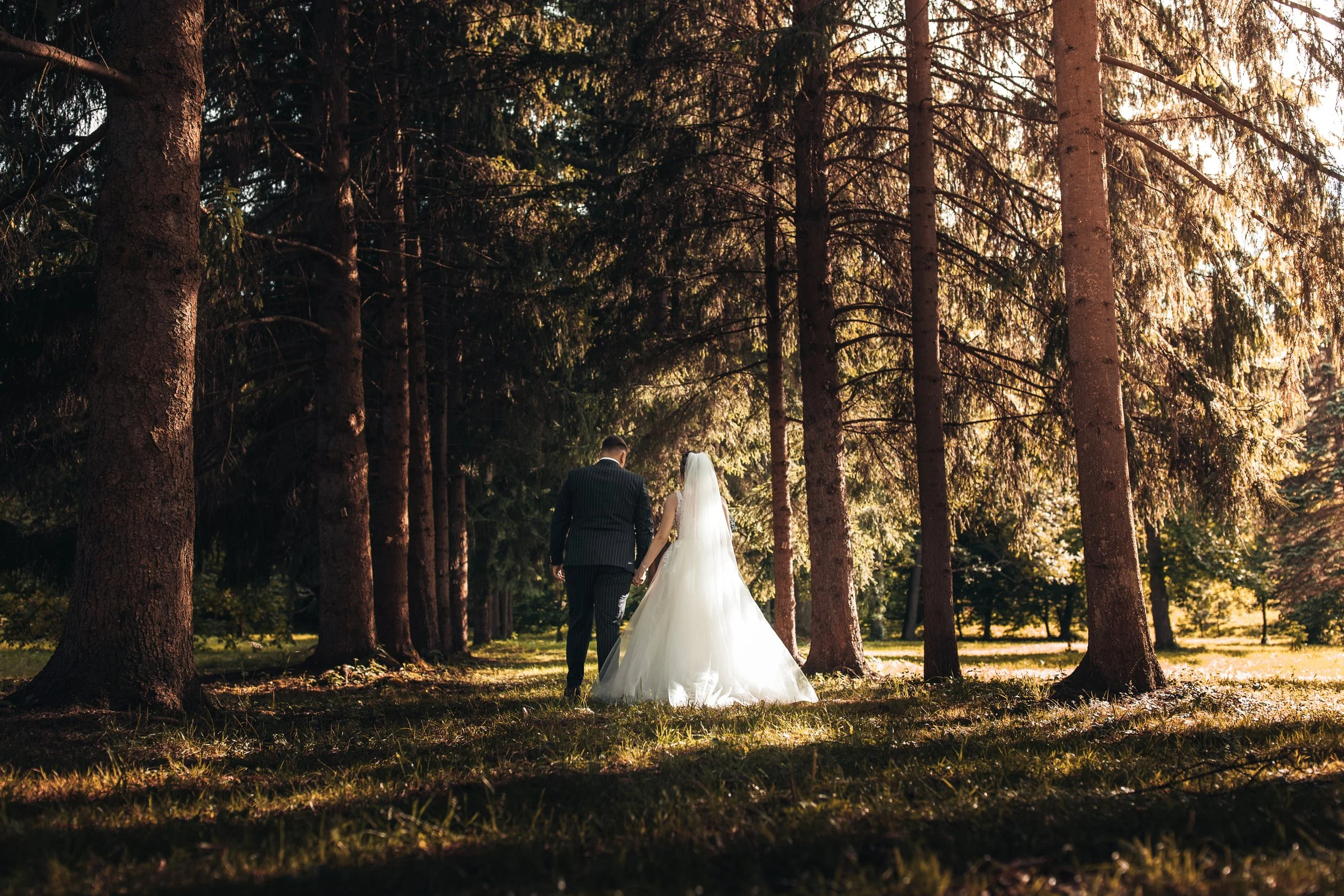 A bride and groom walk hand in hand through a forest with tall trees and sunlight filtering through the branches.