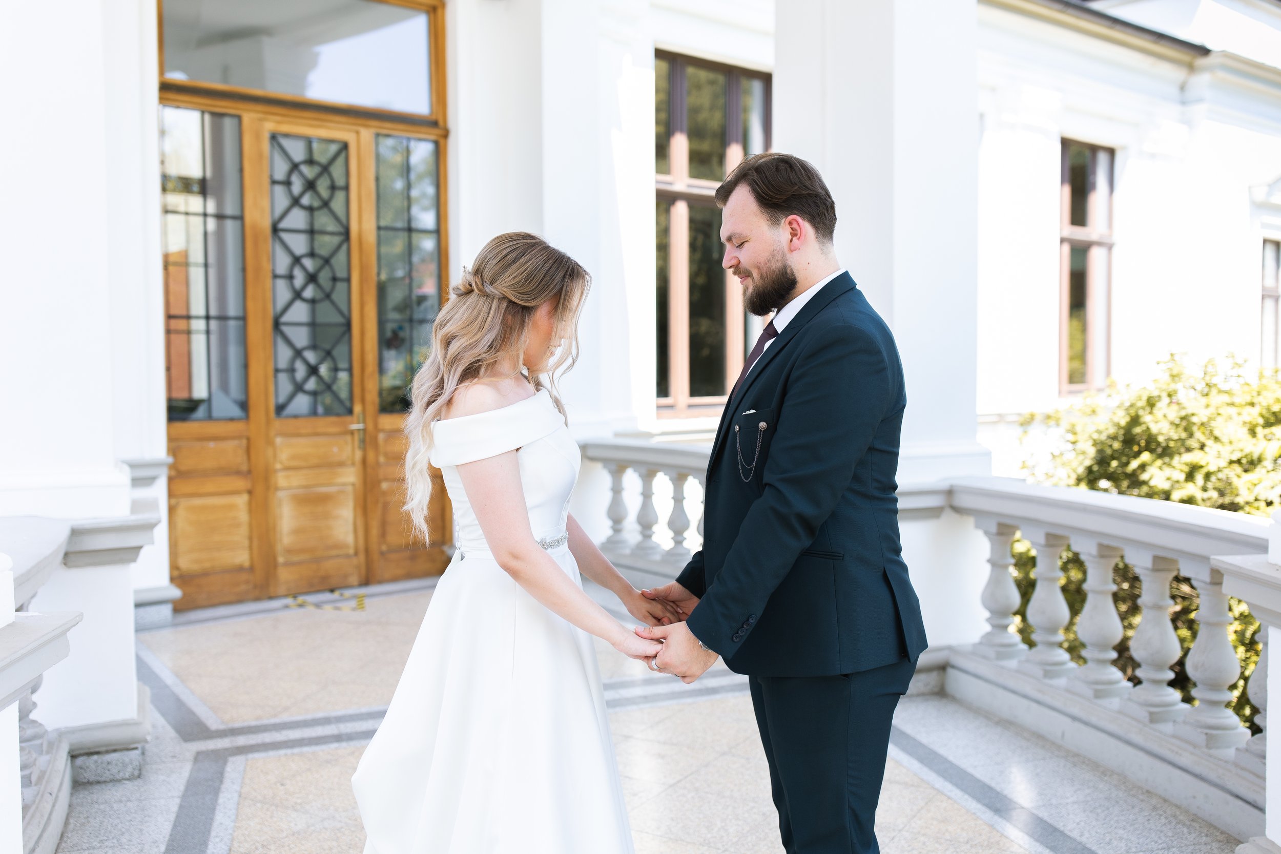 A bride and groom holding hands during their wedding ceremony outside a white building with wooden doors and large windows.