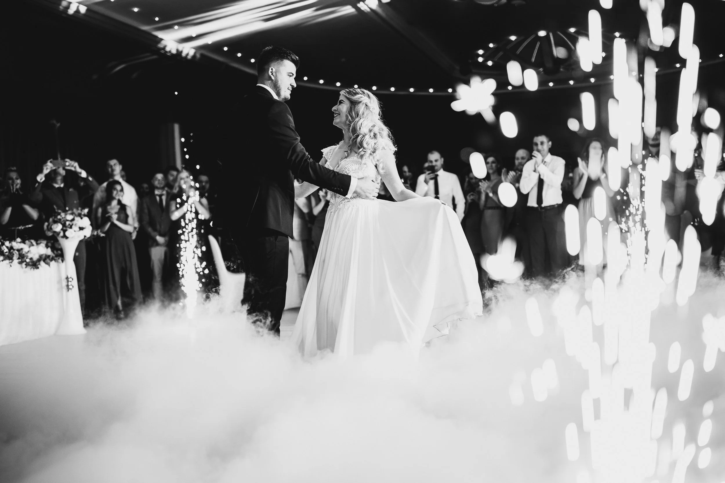 A black-and-white photo of a wedding dance with a bride and groom. The groom is in a suit, and the bride in a wedding gown. They are surrounded by smiling guests, and there are decorative lights, flowers, and fog on the floor.
