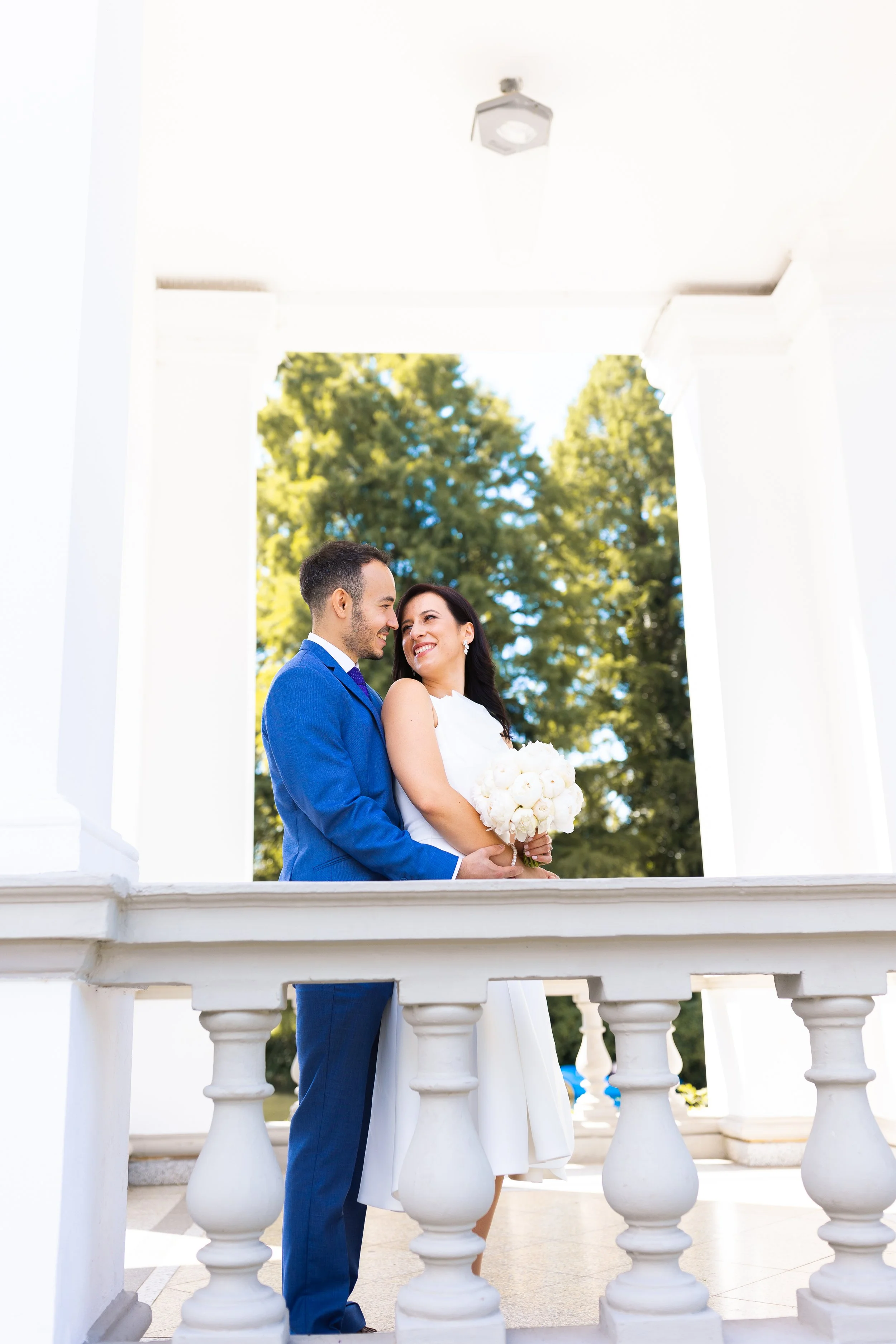 A newlywed couple standing close together and smiling at each other, framed by a white architectural structure with trees in the background. The bride holds a bouquet of white flowers, and the groom is wearing a blue suit.