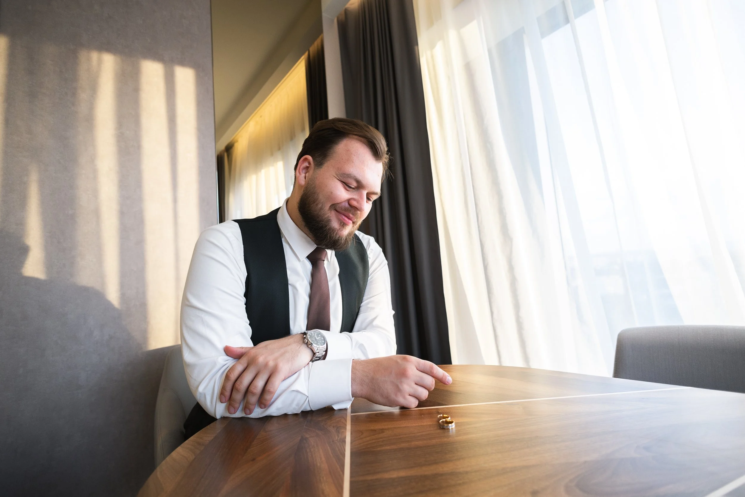 A man in a white shirt, black vest, and brown tie sits at a wooden table, smiling as he looks at two wedding rings placed on the table, with sunlight coming through large windows with white and dark curtains behind him.