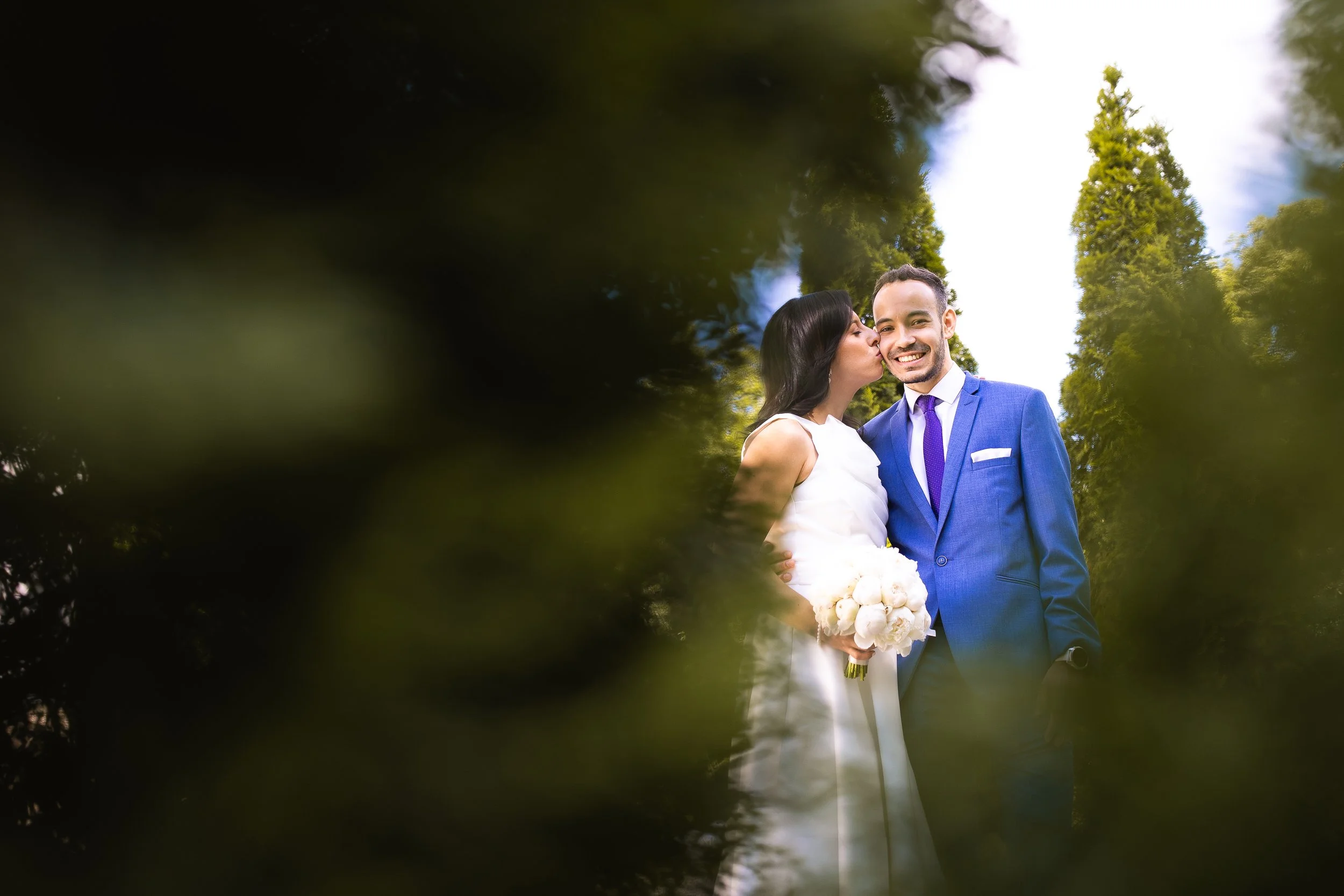 A bride and groom on their wedding day, with the bride kissing the groom on the cheek, standing outdoors among tall trees, with a natural nature backdrop.