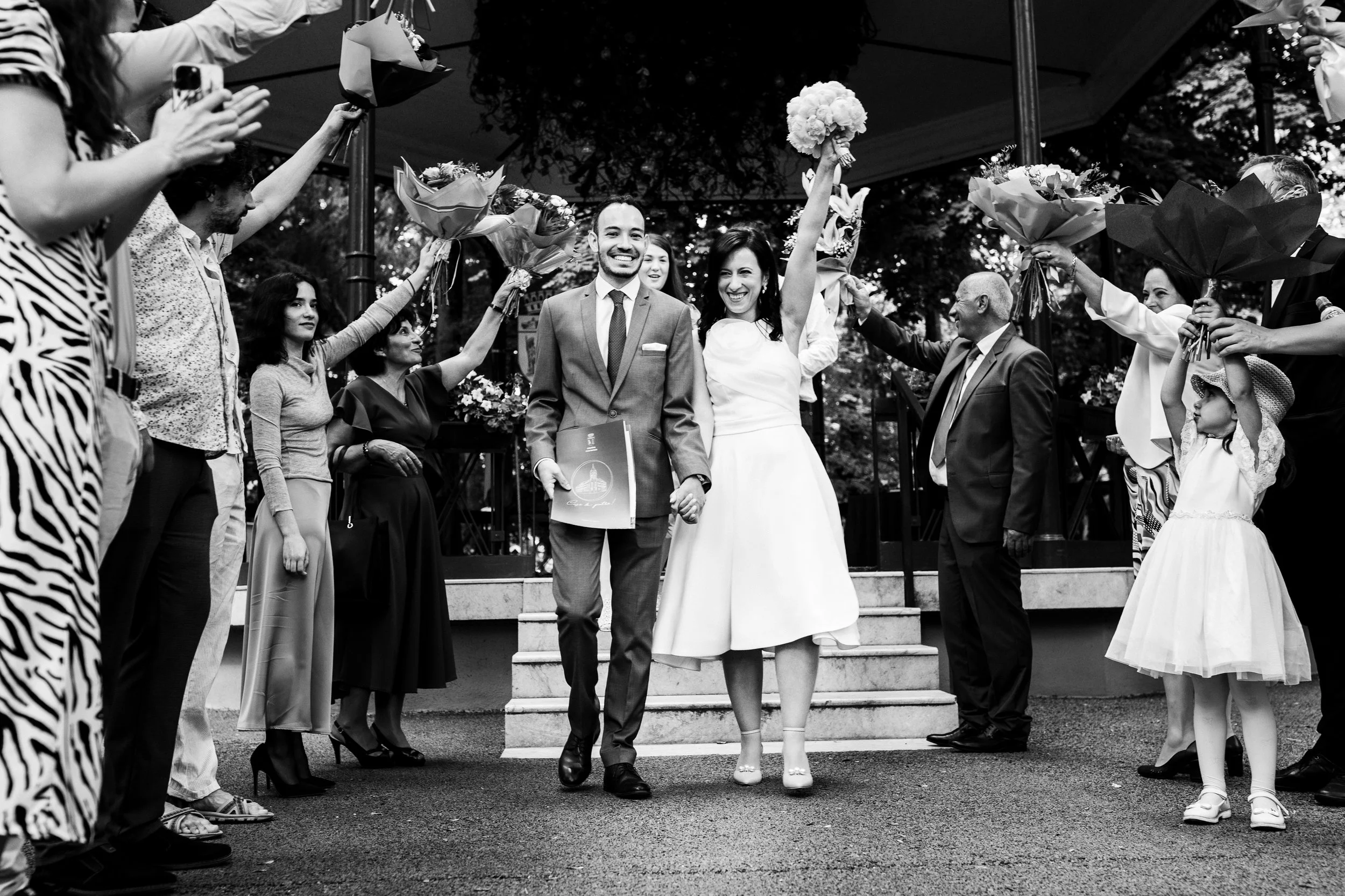 A newlywed couple walking arm in arm, smiling, as they exit a wedding celebration. The bride is holding a bouquet of flowers high above her head, and the groom is holding a certificate or booklet. Friends and family surround them, holding bouquets an