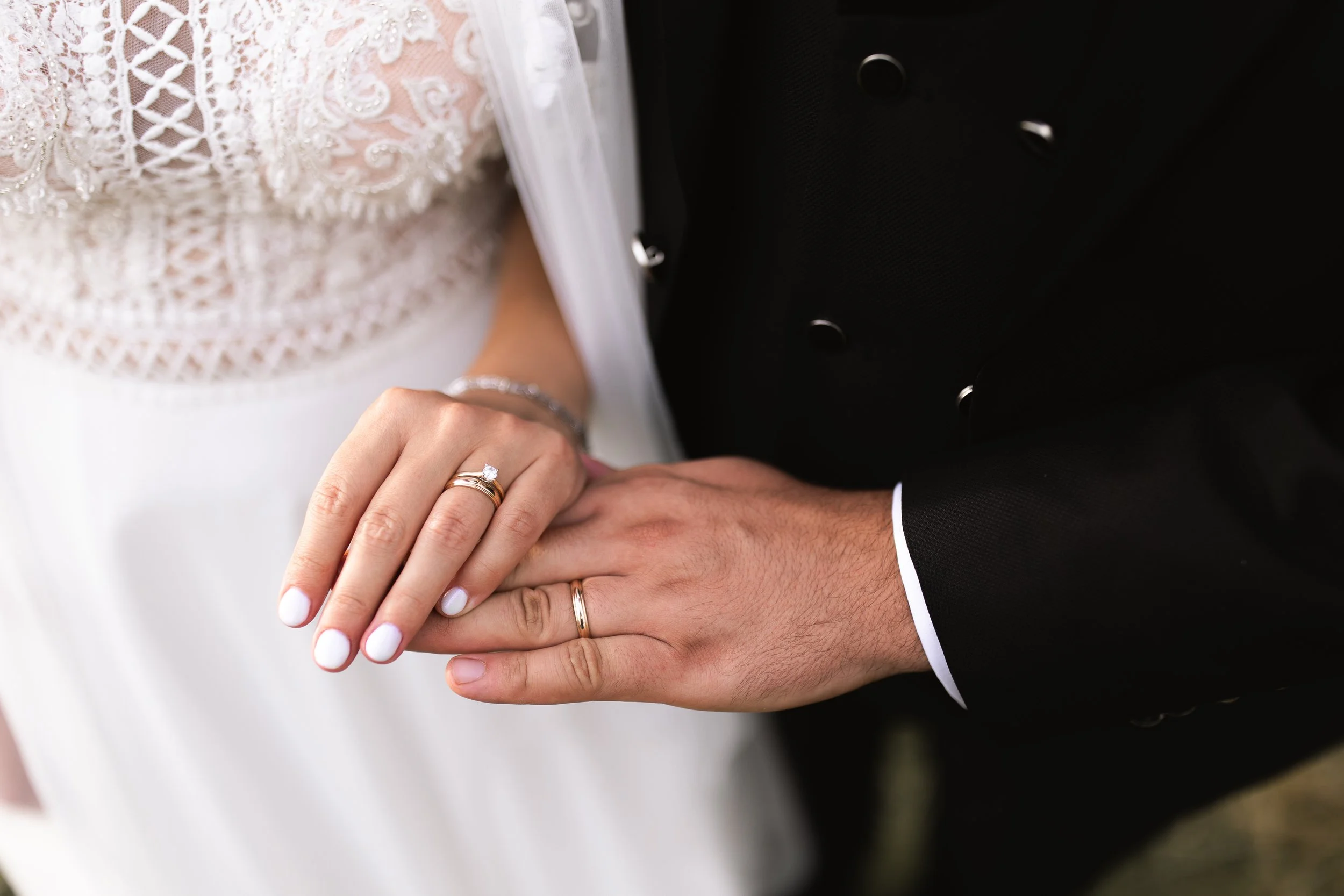 Close-up of a bride and groom holding hands, showing wedding rings. The bride's hand has a diamond engagement ring and a wedding band, with white nail polish. She is wearing a bracelet. The groom's hand has a plain wedding band. The bride is wearing 