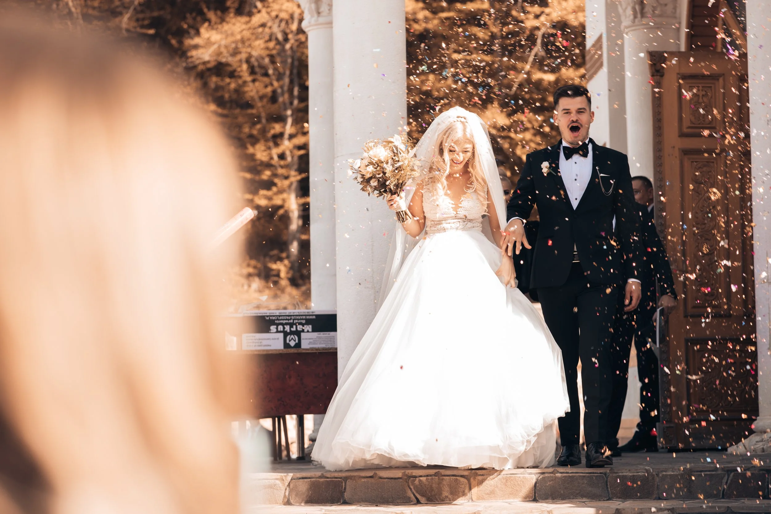 A bride and groom walking out of a building after their wedding, surrounded by falling confetti, with the bride holding a bouquet of flowers and smiling.