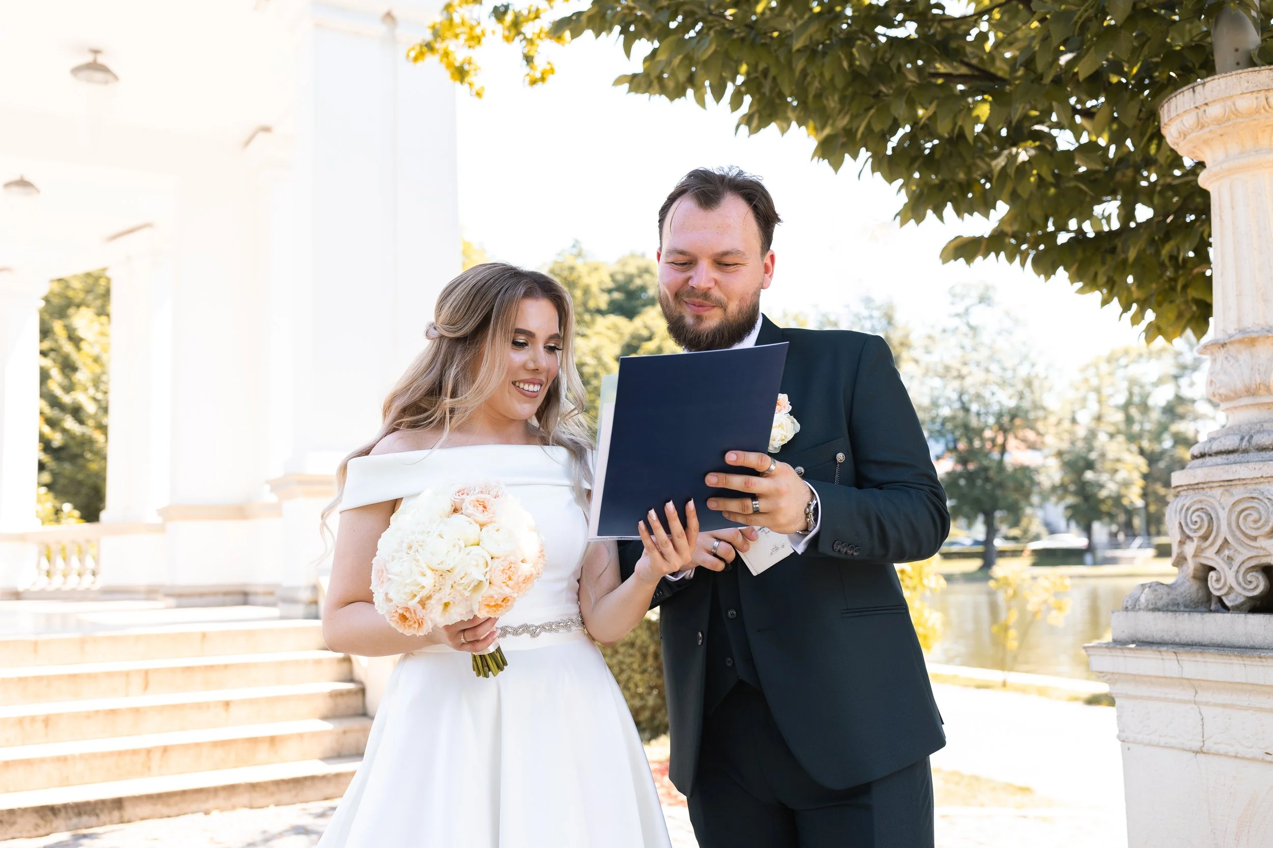 A bride in a white wedding dress holding a bouquet and a groom in a black suit reading a document outdoors during their wedding ceremony.