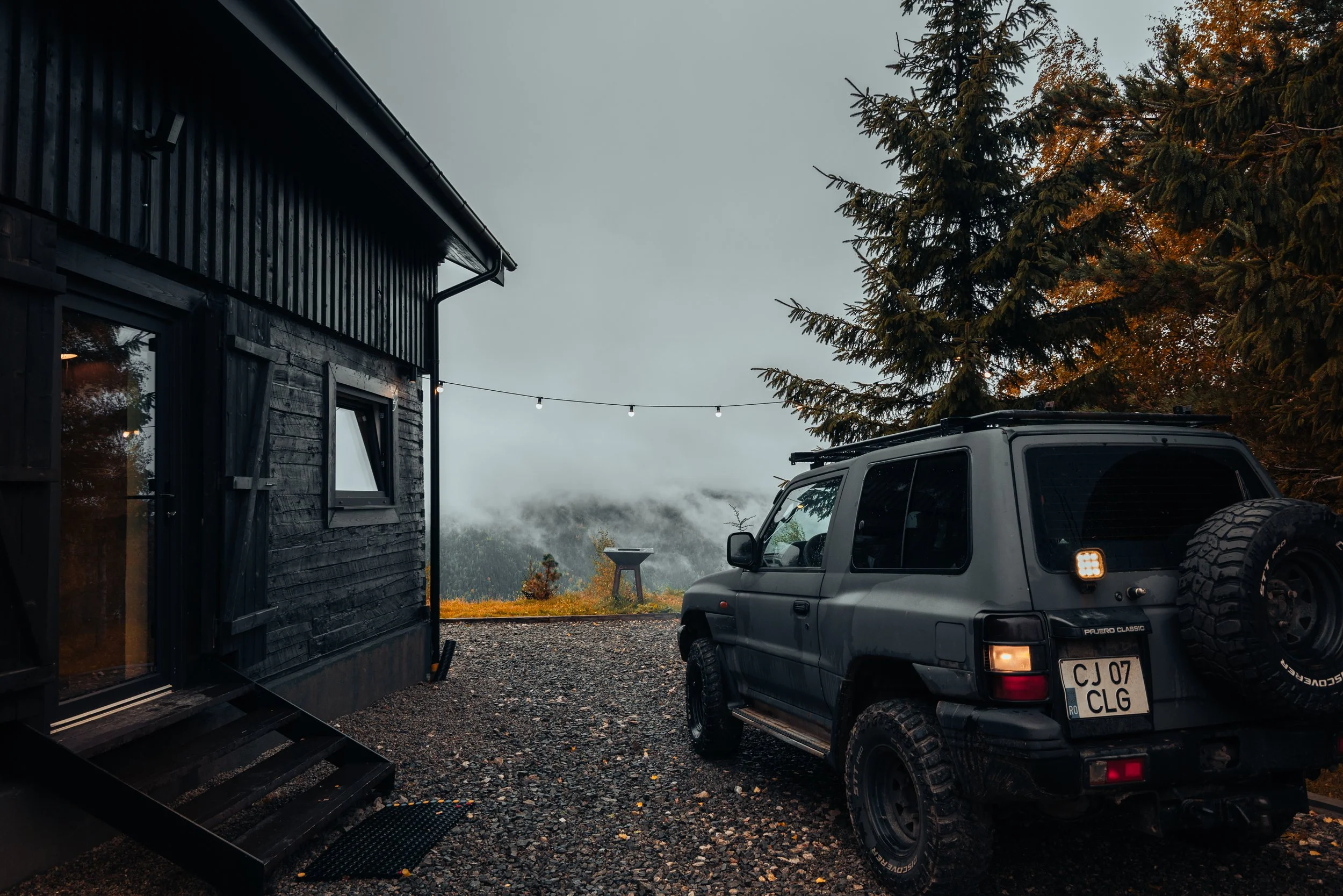 A black wooden cabin and a gray SUV parked outside on a gravel surface in a foggy forest setting with pine trees and string lights overhead.