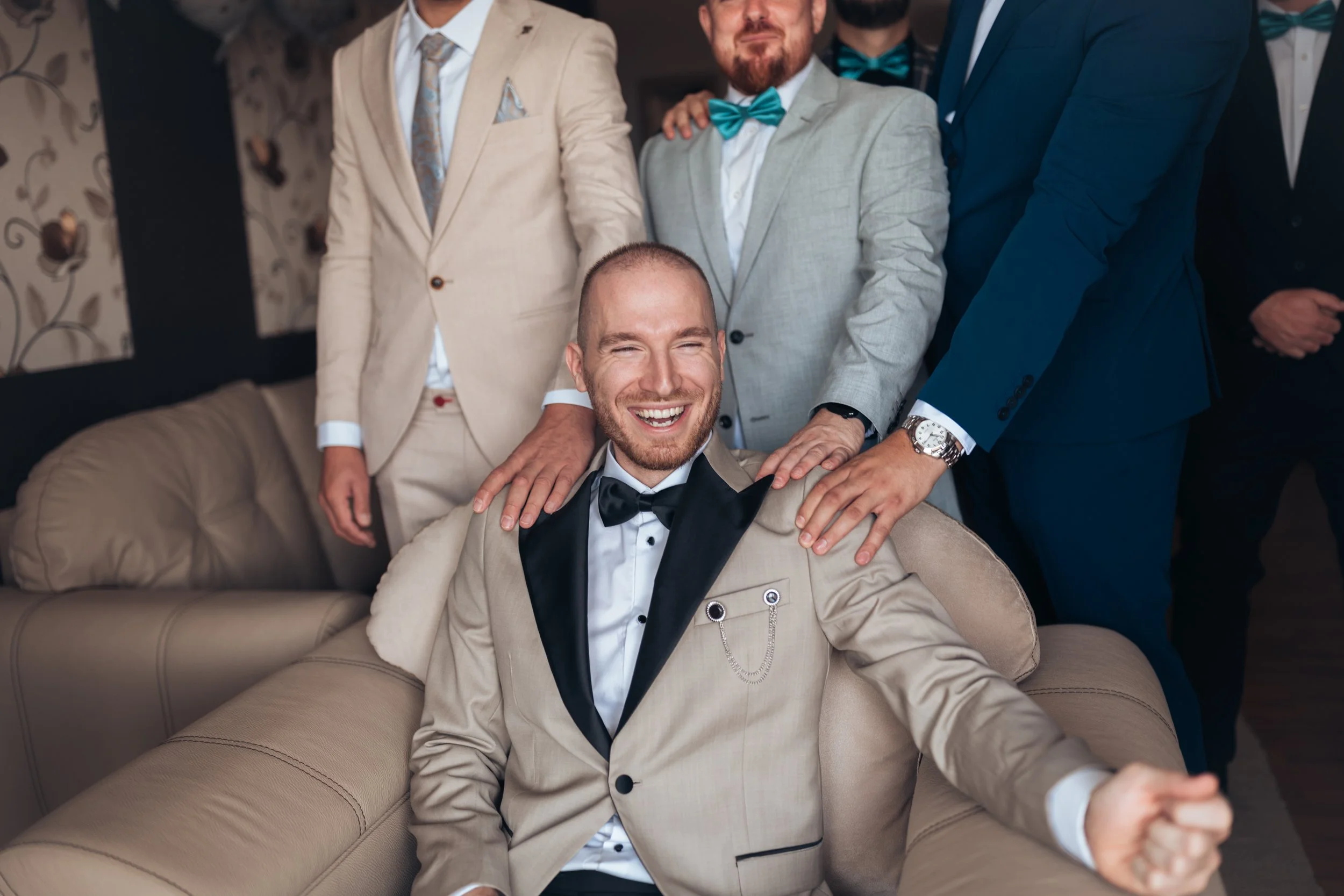 A group of men in suits and tuxedos gathered around a man sitting on a beige sofa, smiling with a joyful expression, during a formal occasion.