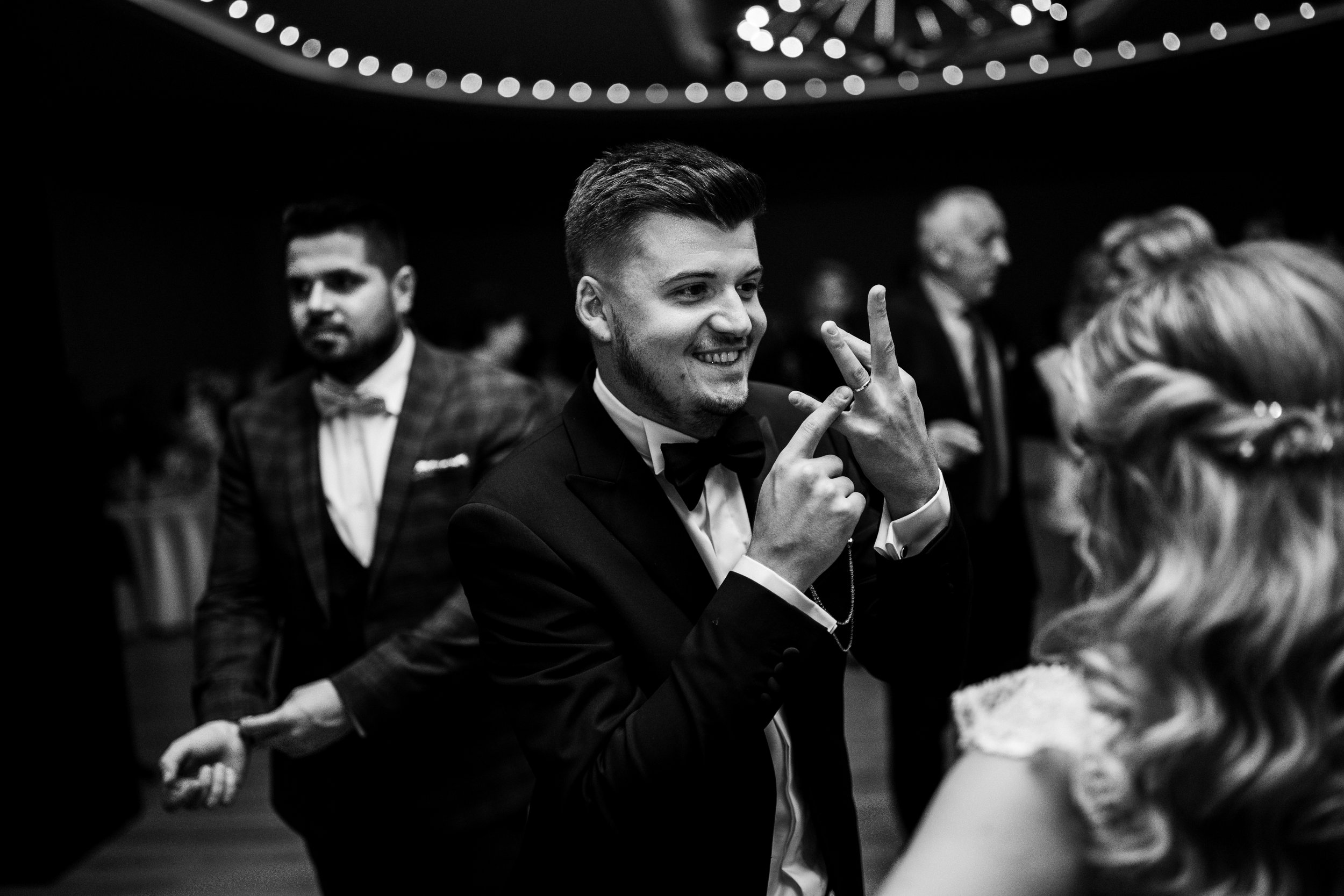 A young man in a tuxedo making a peace sign while dancing at a formal event, with other guests in the background.