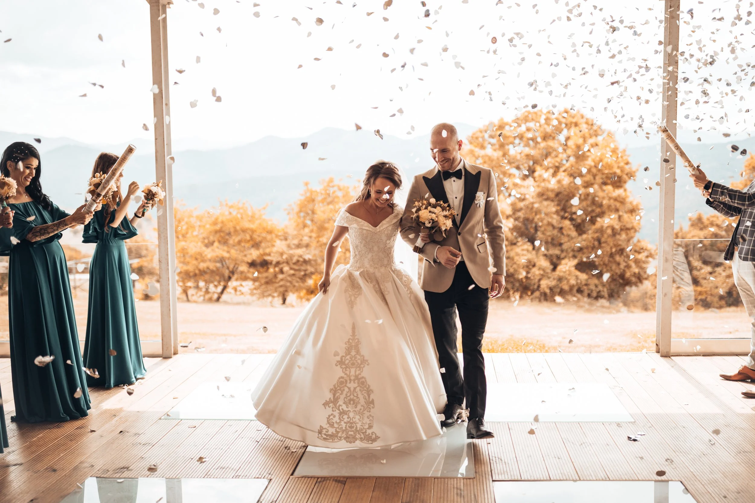 Bride and groom walking down the aisle under falling confetti during their outdoor wedding ceremony, with bridesmaids in teal dresses on the left and groomsmen on the right, and scenic trees and mountains in the background.