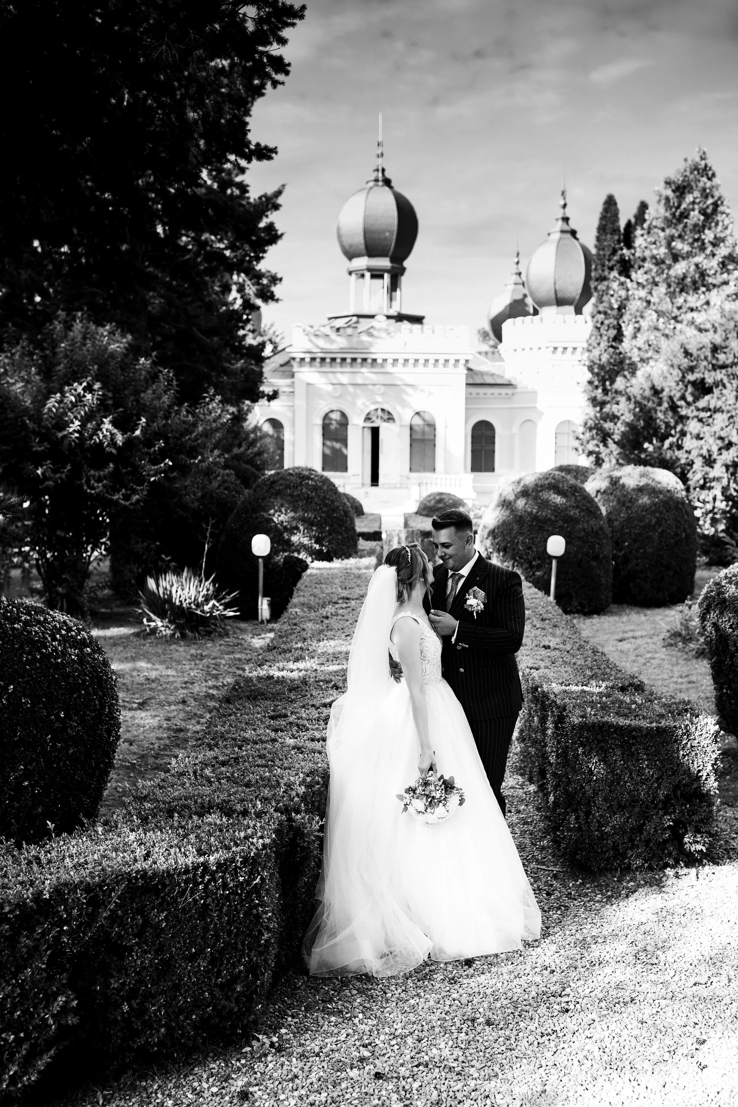 A black and white photo of a bride and groom standing together outdoors in a garden, with a castle-like building with onion domes in the background. The bride is holding a bouquet and wearing a wedding dress and veil, while the groom is in a dark sui