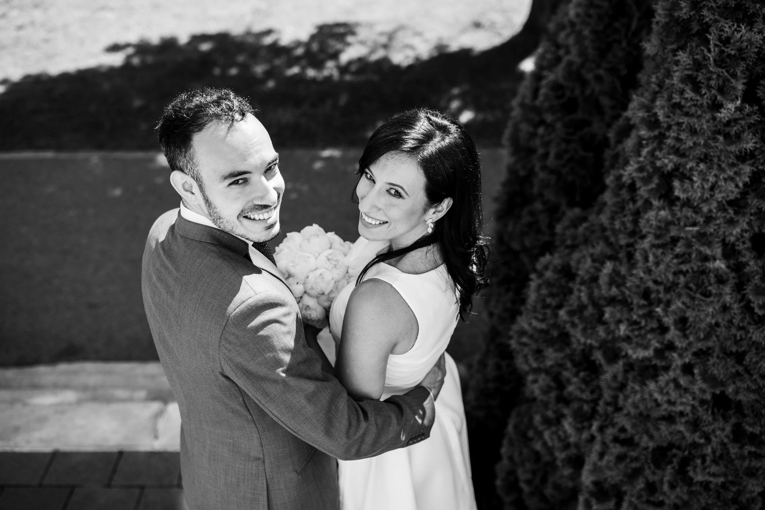 Black and white photograph of a smiling bride and groom embracing outdoors, with trees and a scenic landscape in the background.