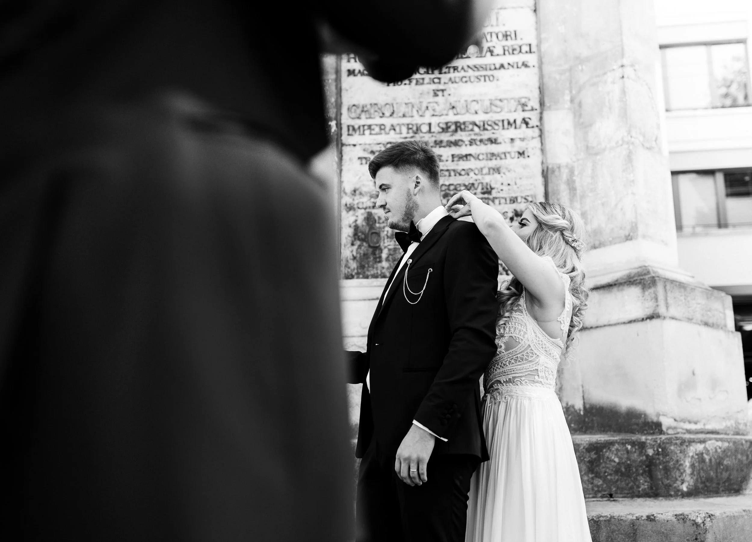 A bride adjusting the tuxedo of the groom in front of a historical building with engraved text, during a wedding photoshoot in black and white.