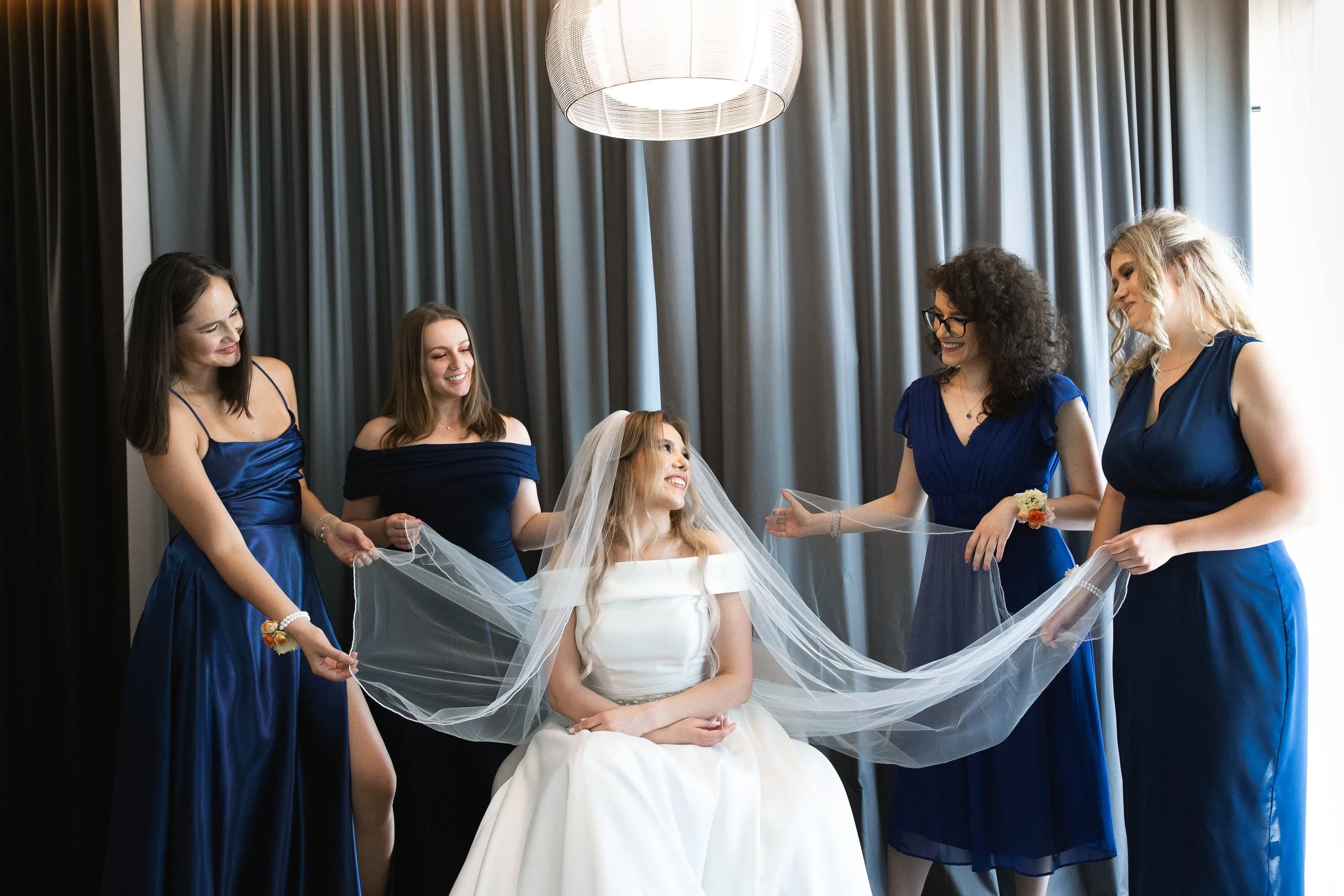 Bride in a white dress sitting and smiling, surrounded by five women in matching blue dresses, holding her veil in a well-lit room with dark curtains.