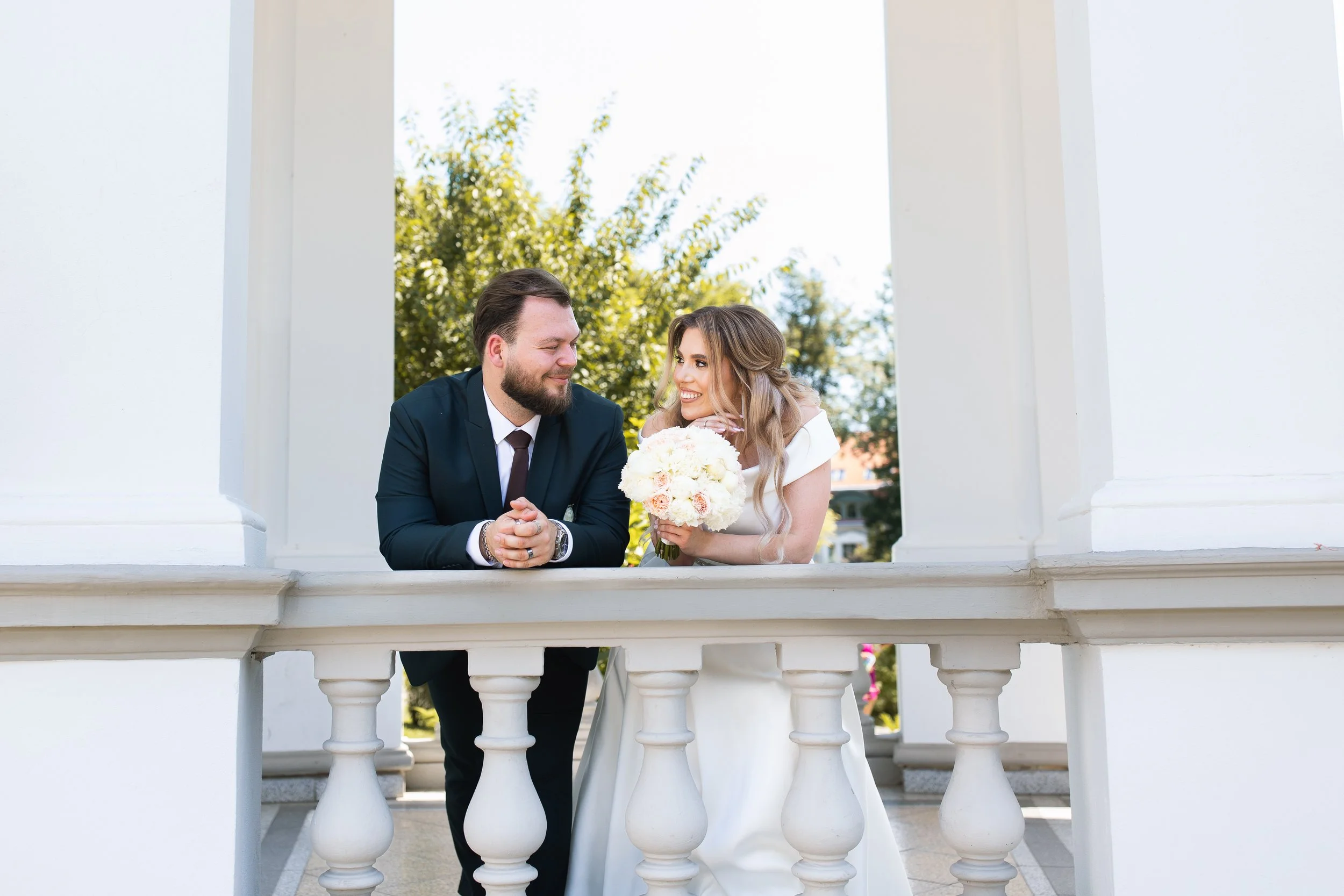 A newlywed couple leaning on a white balcony railing, smiling at each other. The bride holds a bouquet of white and pink roses. They are outdoors with green trees in the background.