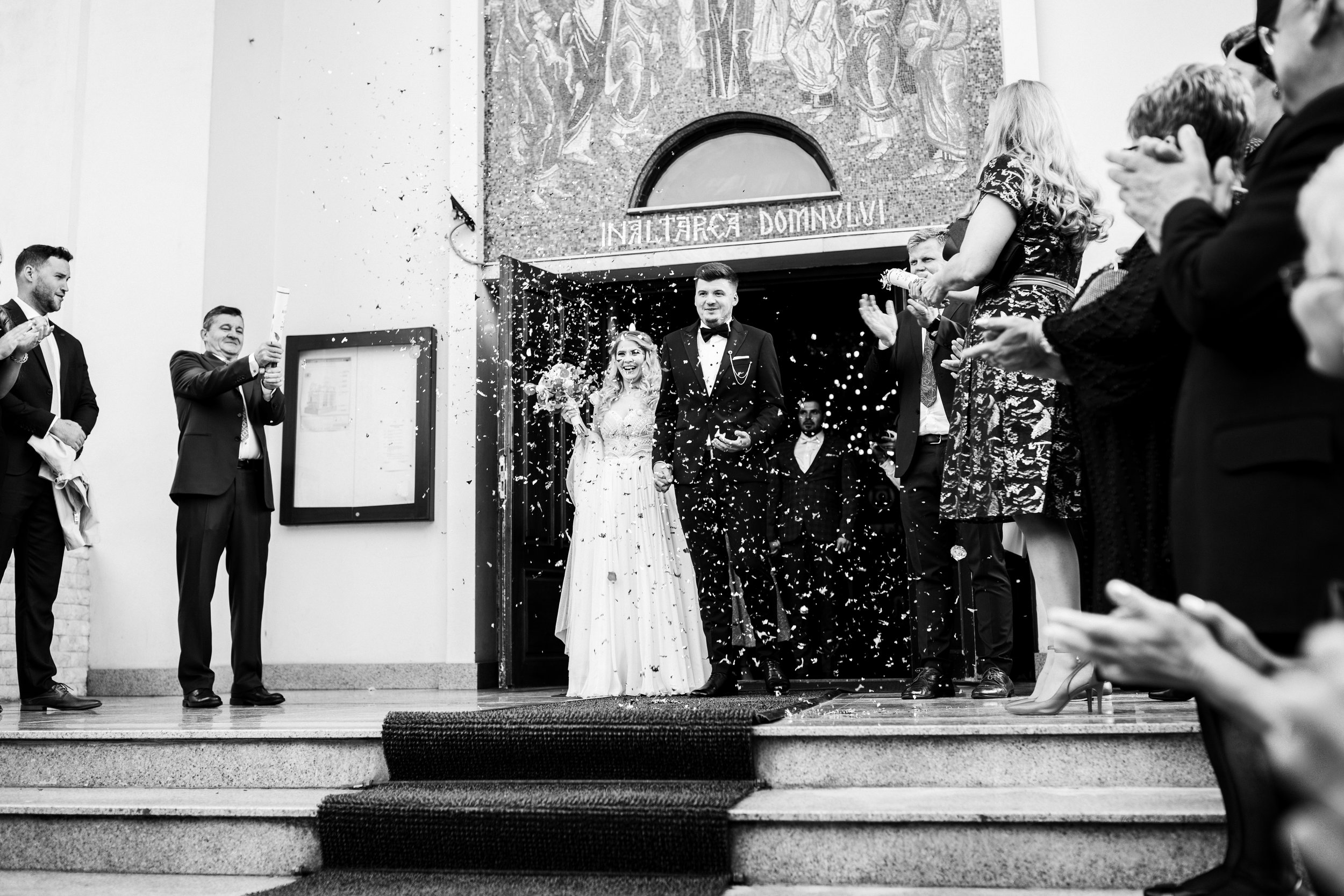 Black and white photo of a bride and groom leaving a church, surrounded by guests holding and throwing confetti and clapping, with a church entrance and mural in the background.