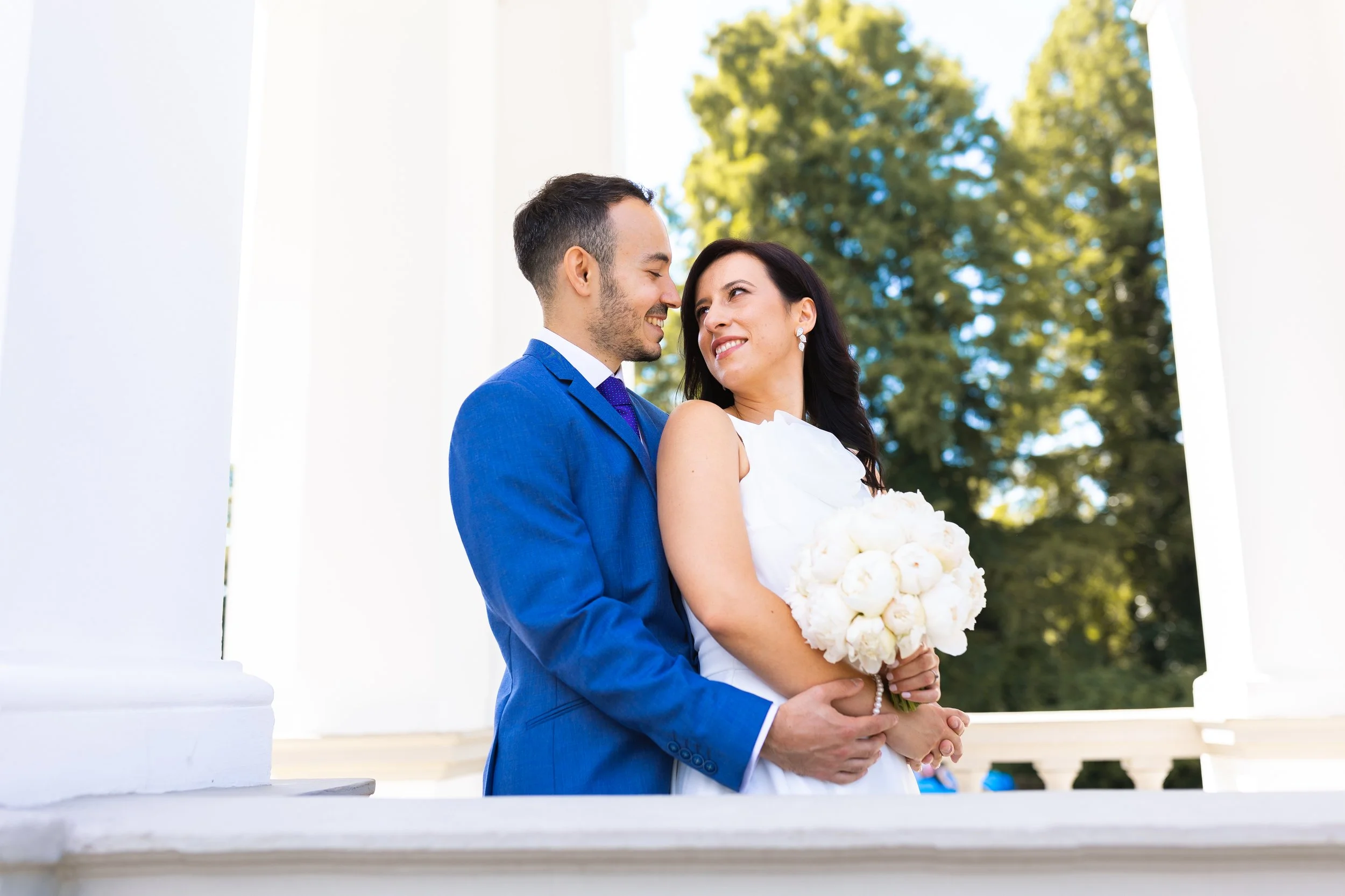 A newlywed couple, the groom in a blue suit and the bride in a white dress holding a bouquet of white flowers, embracing lovingly outdoors with green trees in the background.