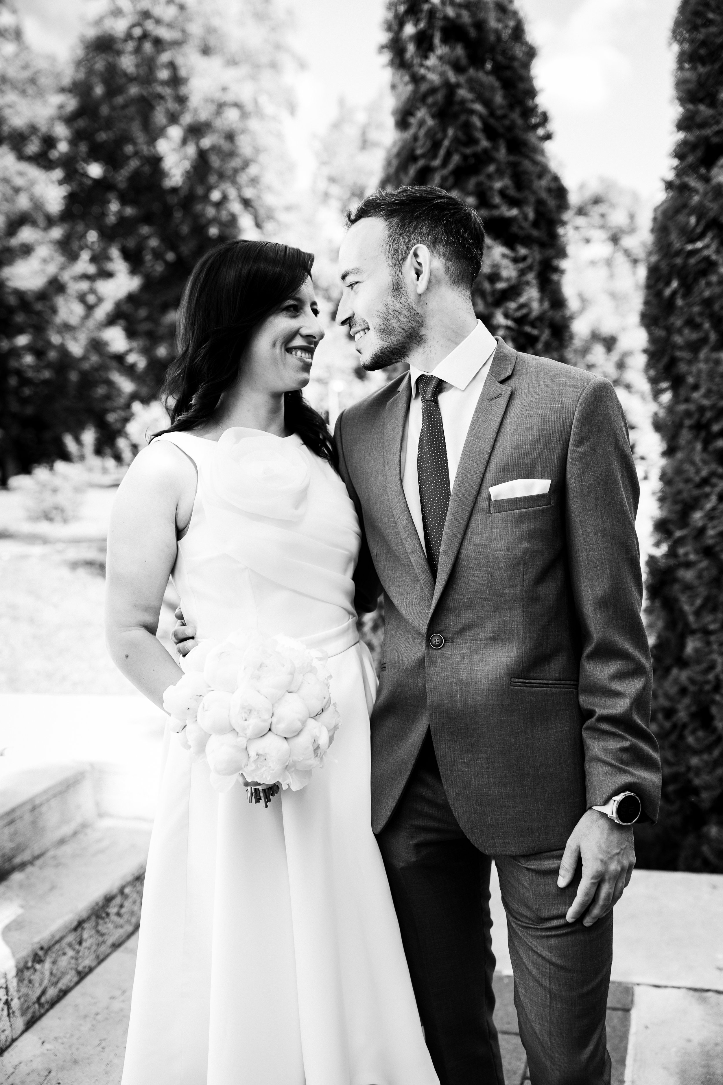 A black-and-white photo of a bride and groom standing close together outdoors, smiling and gazing into each other's eyes, with trees in the background. The bride holds a bouquet of flowers, and the groom is dressed in a suit with a watch on his wrist