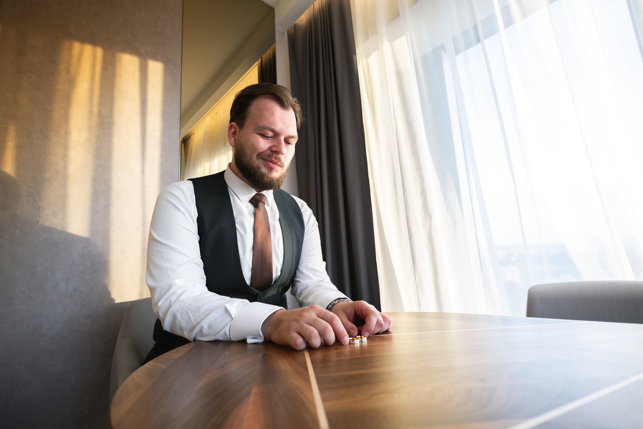 A man in a white shirt, black vest, and brown tie sitting at a wooden table, smiling and organizing gold coins, near large windows with white and dark curtains.