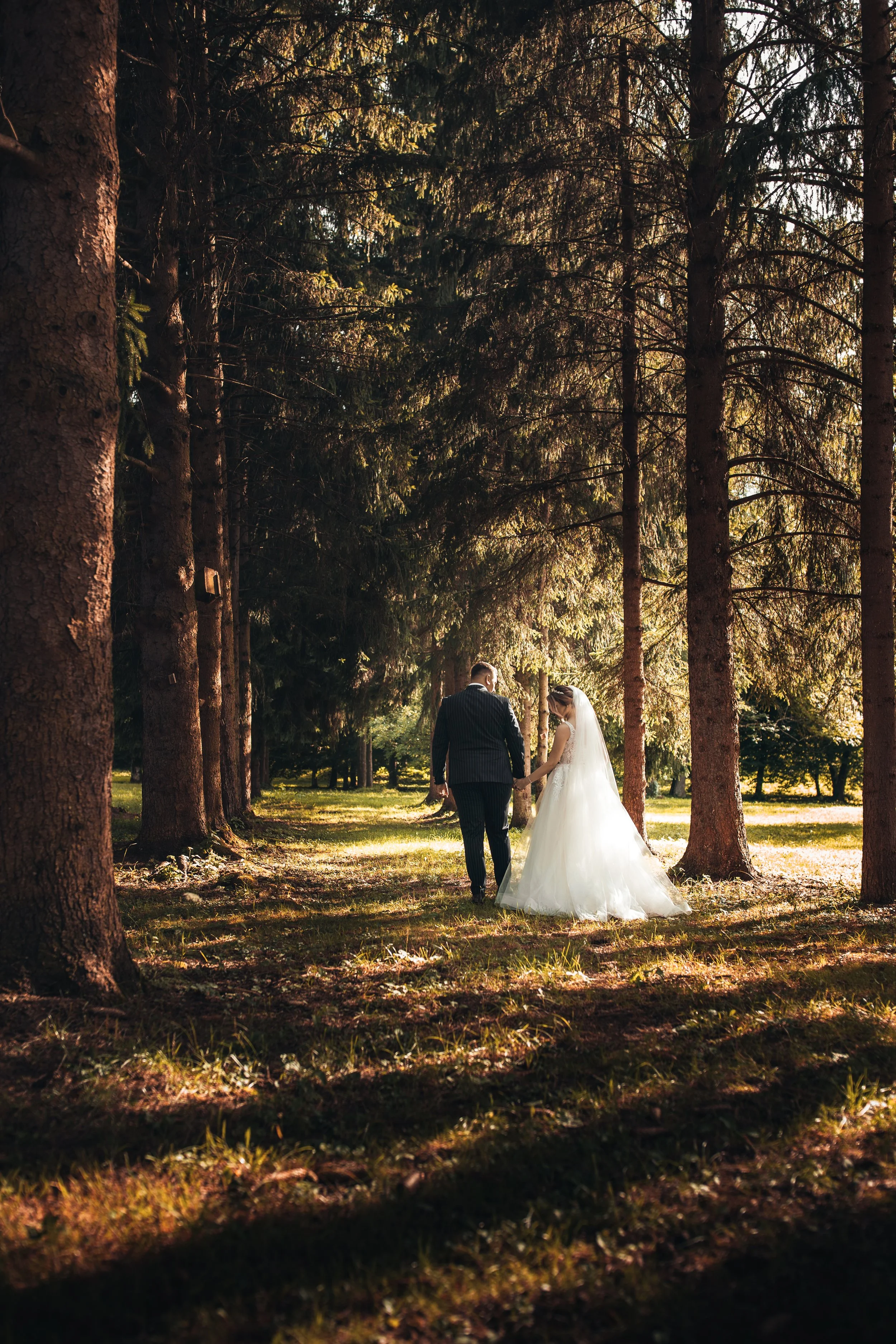 A bride and groom walk hand in hand through a wooded area during their wedding, surrounded by tall trees and sunlight filtering through the branches.
