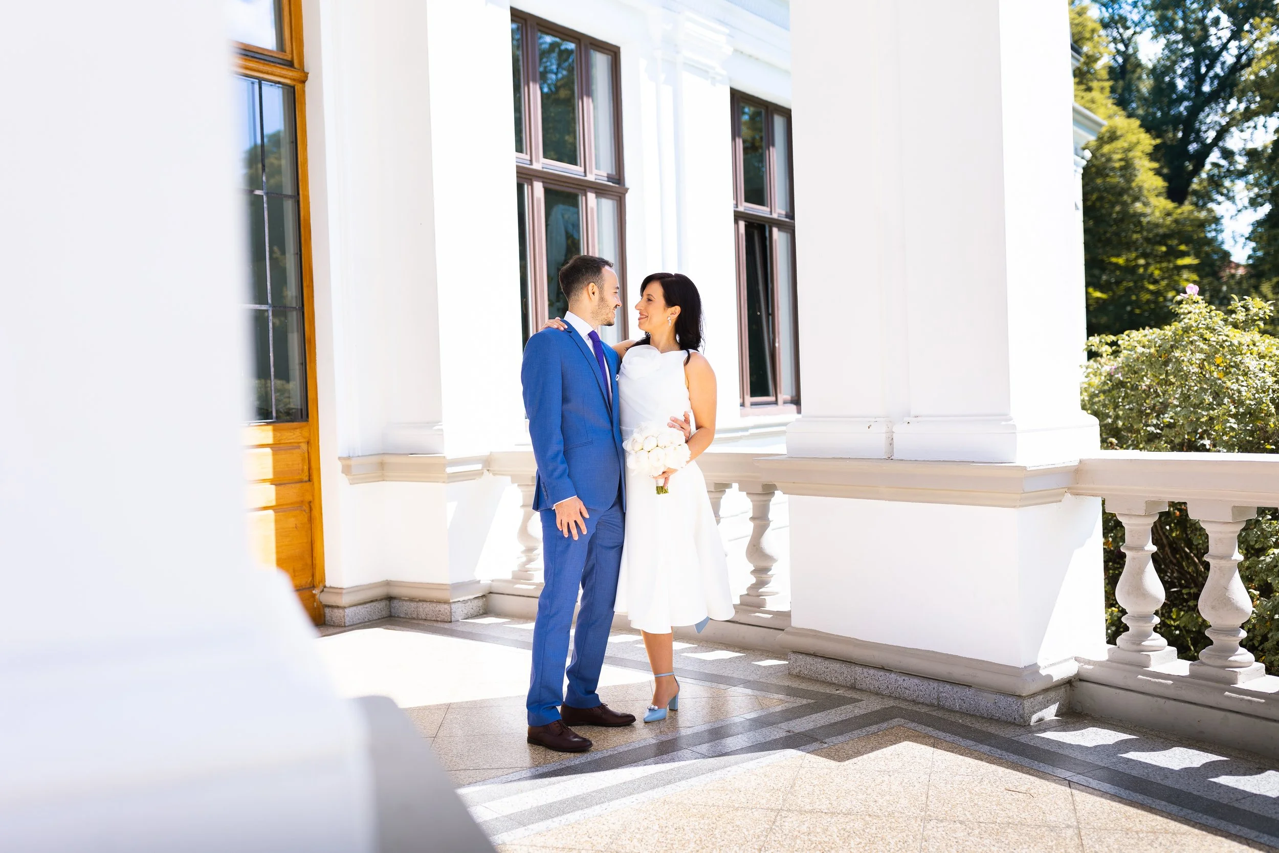 A couple on a balcony, dressed in wedding attire, smiling and looking at each other. The man is wearing a blue suit, and the woman is wearing a white dress holding a white bouquet. There are white columns and large windows in the background, with tre