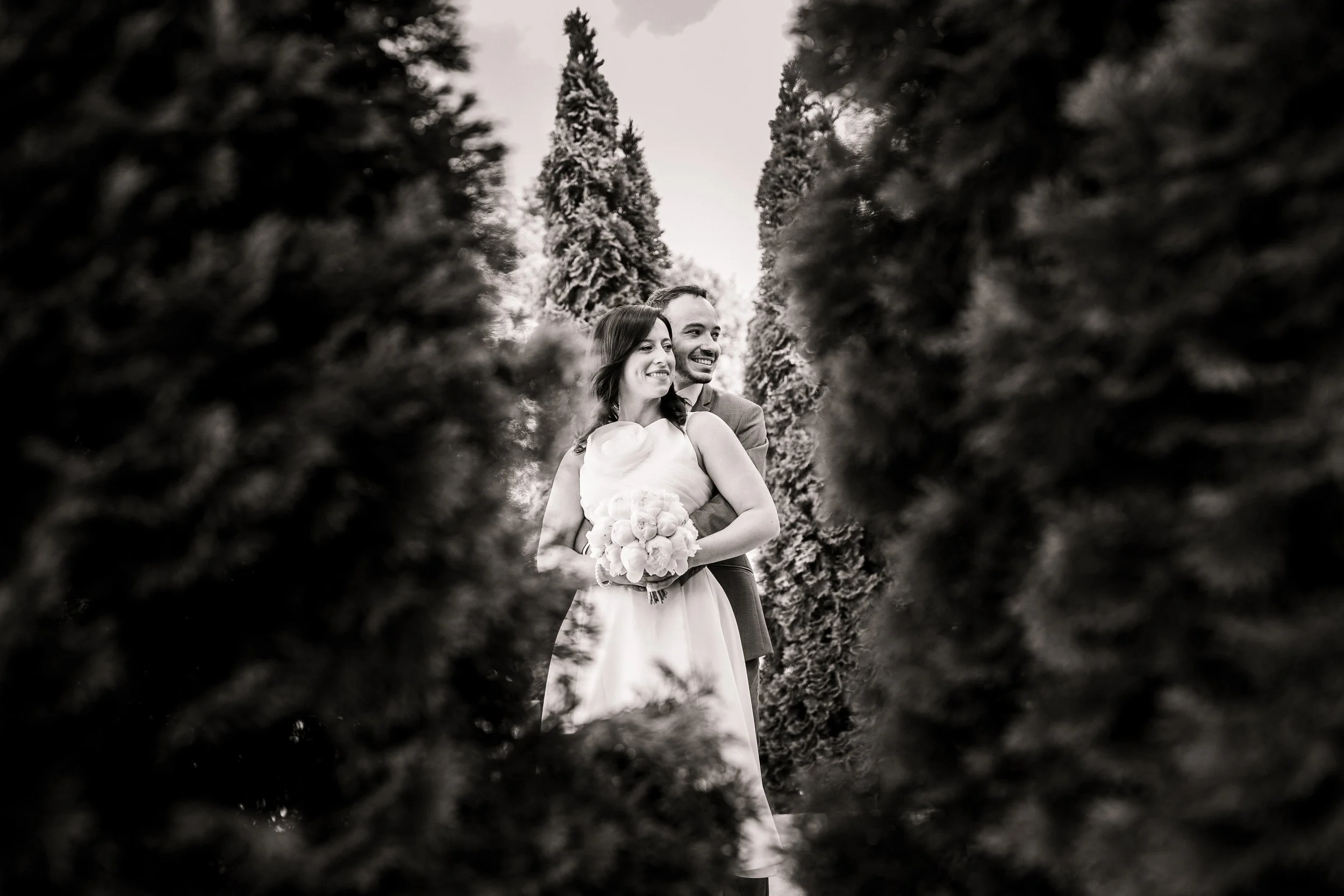 A black and white photograph of a happy couple on their wedding day, embracing and smiling, with the woman holding a bouquet of flowers, surrounded by tall trees, seen through a gap in shrubbery.