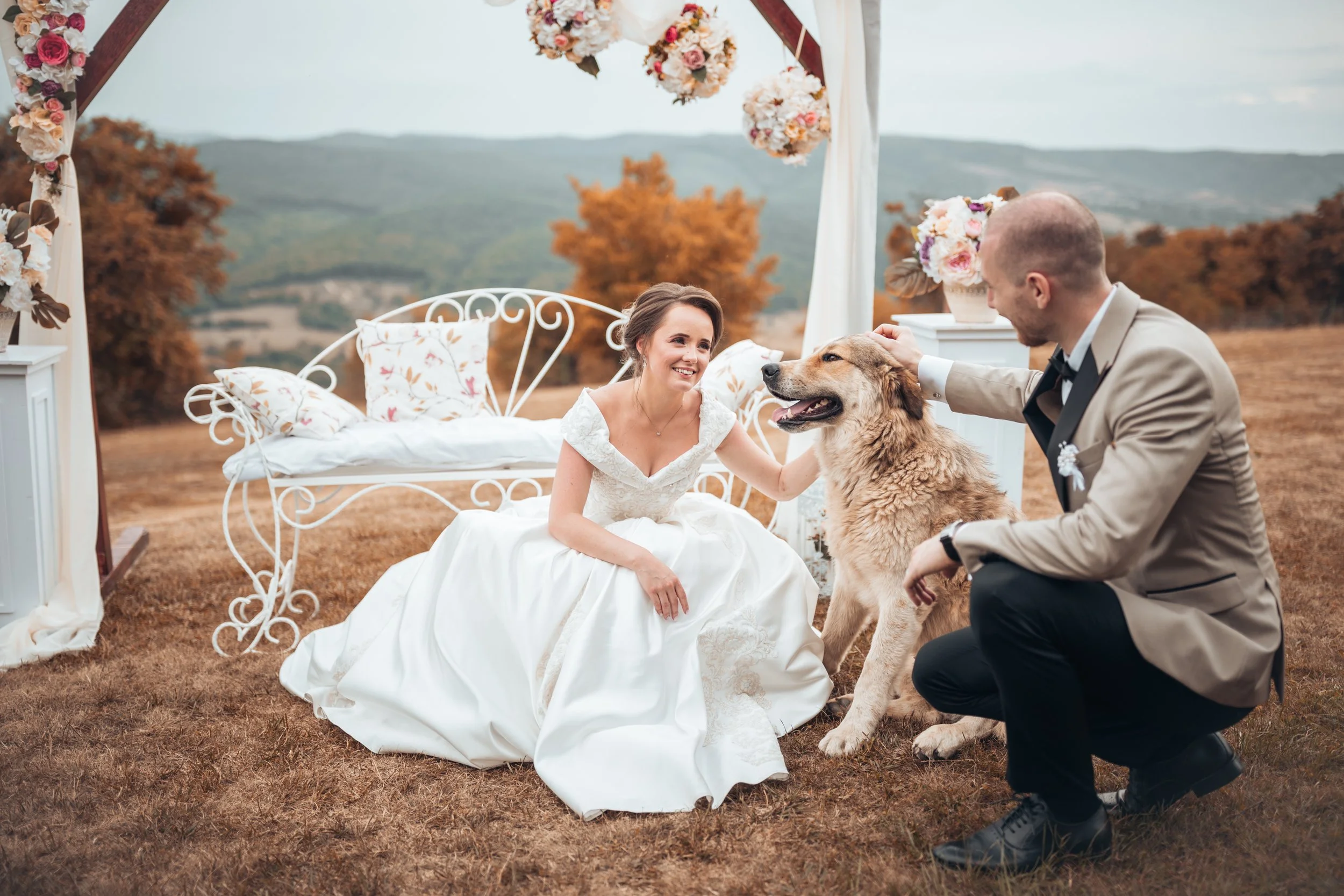 A bride and groom outdoors at their wedding, with the bride sitting on the ground in a white wedding dress and the groom kneeling beside her, petting a large dog. The setting includes a white wrought iron bench and floral decorations, with mountains 