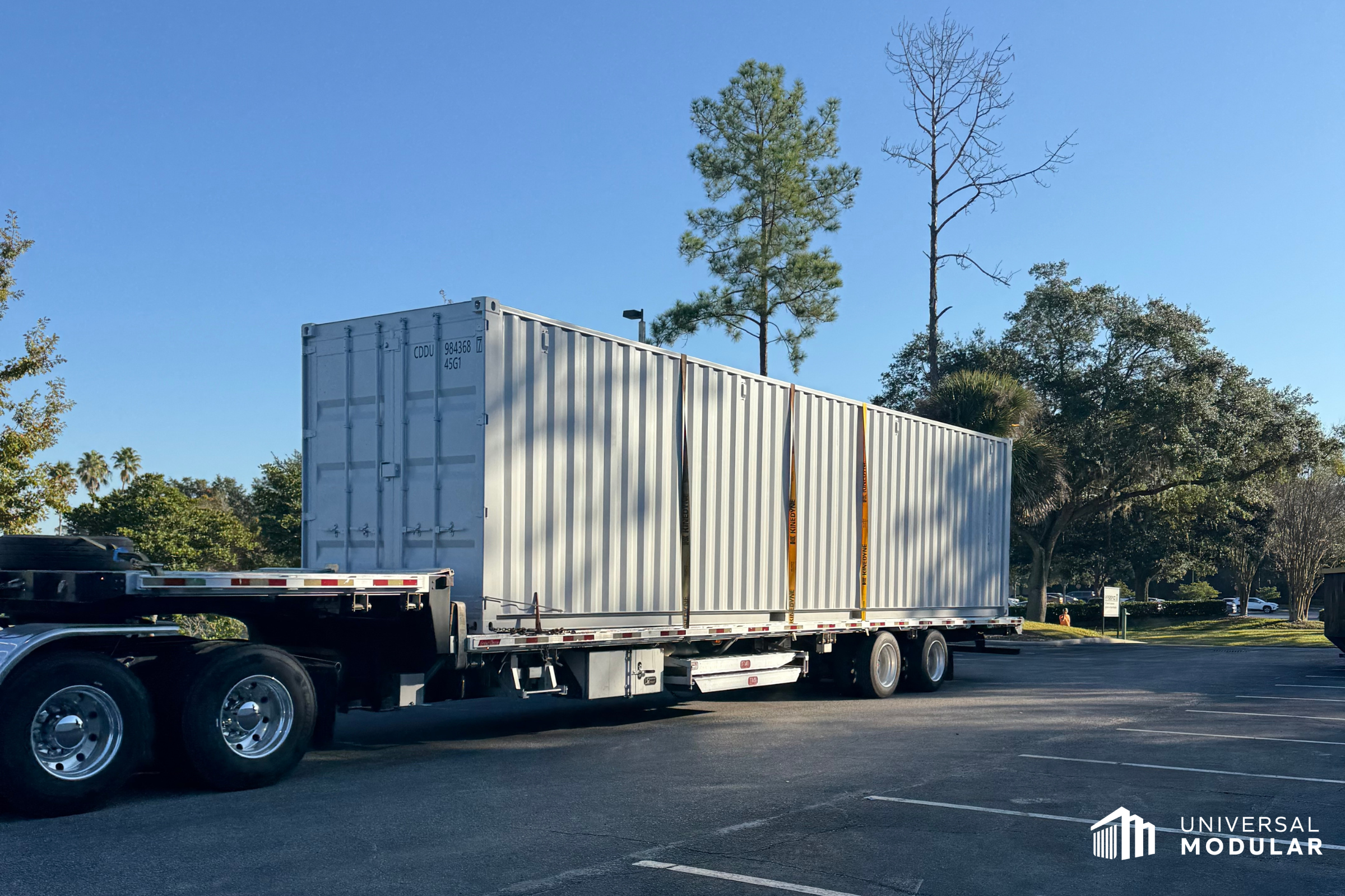 container scif, aka scif in a box, is strapped to truck for delivery in asphalt parking lot with trees and blue sky in background