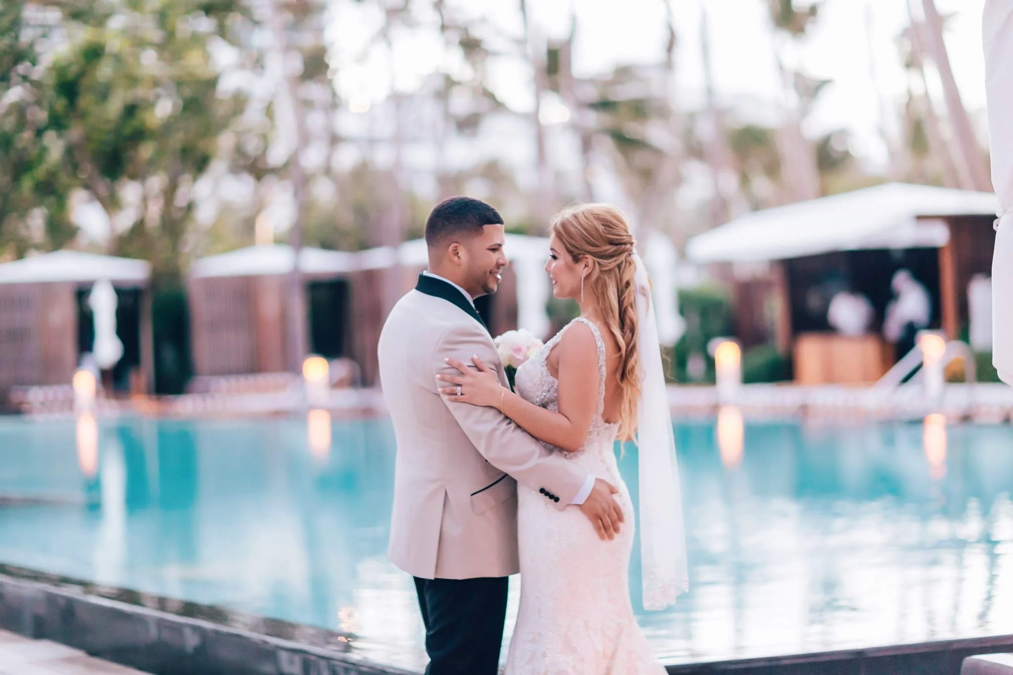A bride and groom smiling and embracing near a pool during their wedding celebration, with trees and outdoor furniture in the background.