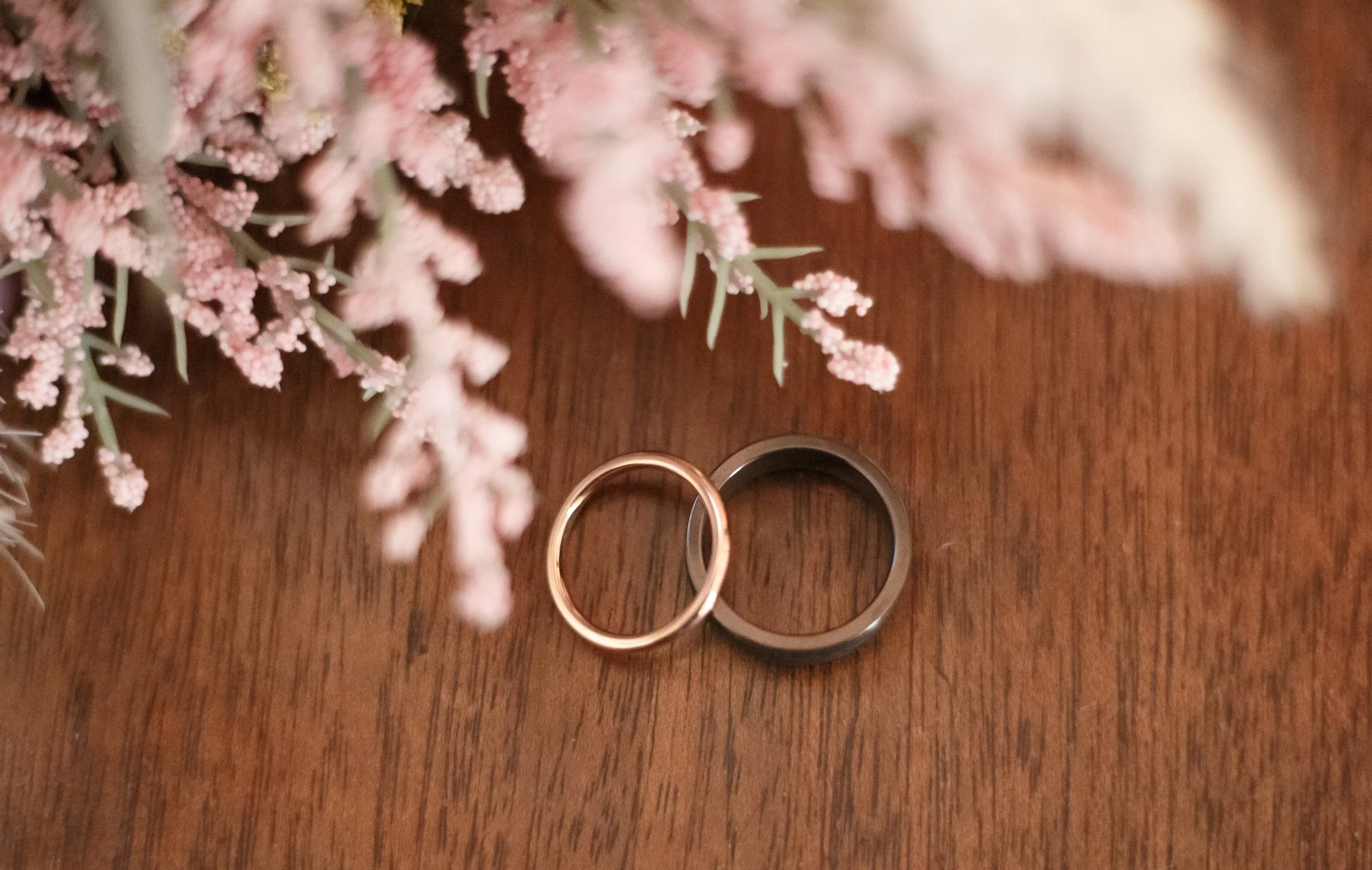 Two wedding rings, one gold and one black, on a wooden surface with pink floral arrangement above.