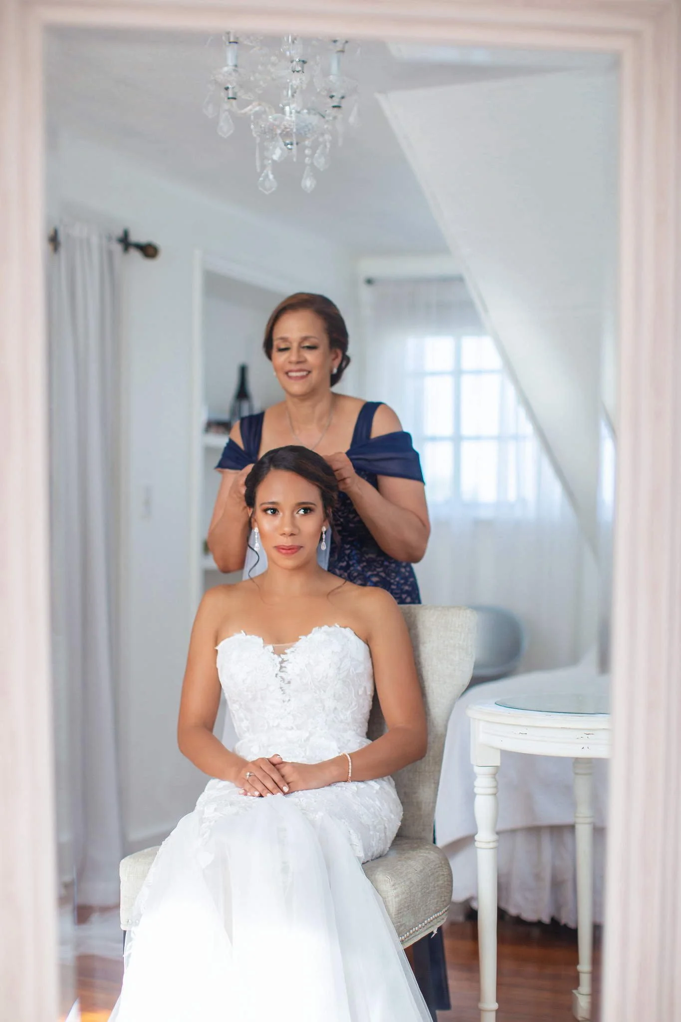 A bride in a white wedding dress sitting on a chair while a woman styles her hair in a room with natural light and a chandelier.