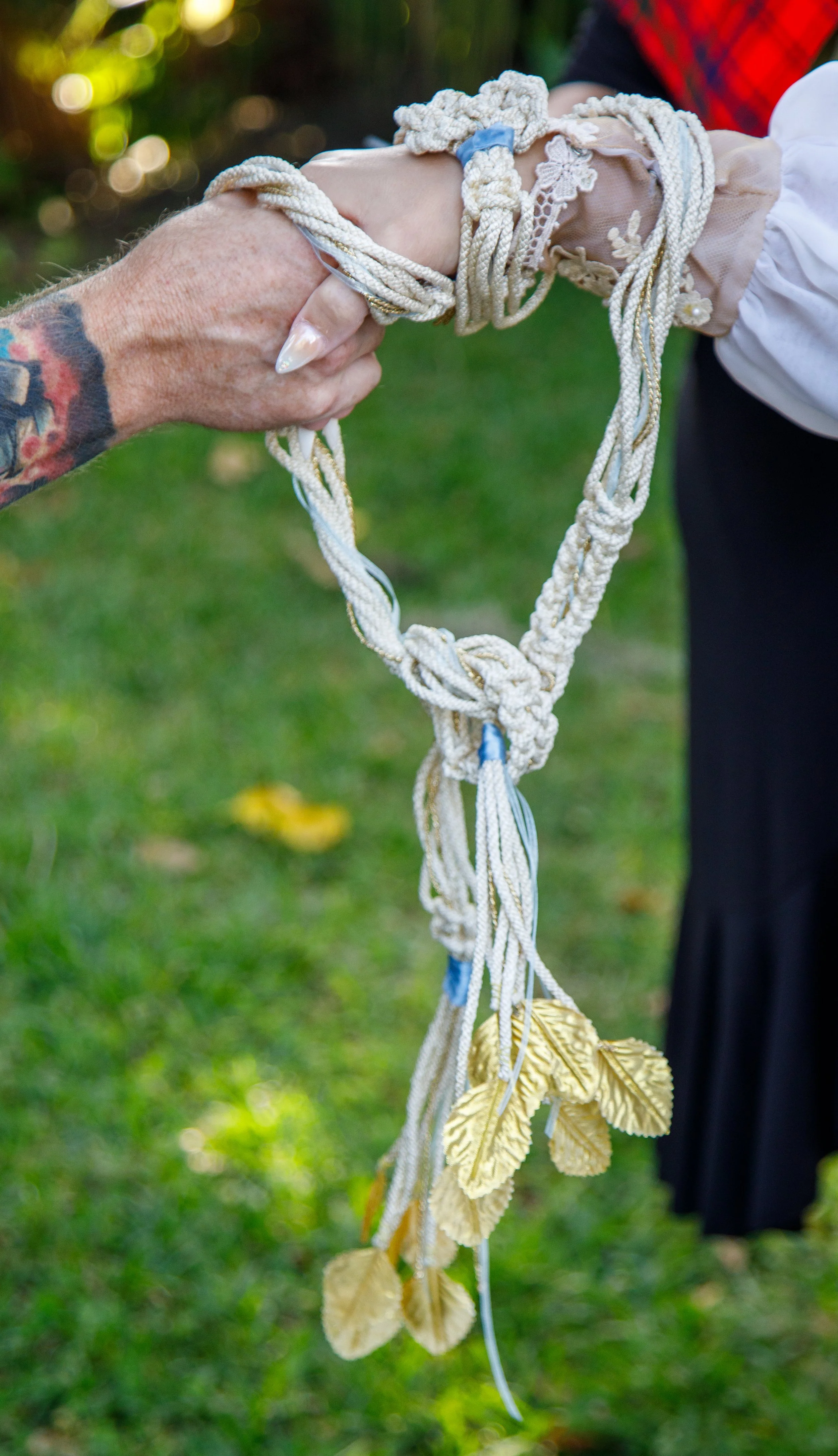 Two people hold a heart-shaped object made from intertwined white rope with gold leaves hanging from its bottom. One person has a tattooed arm, and the other is wearing a lace and embroidered sleeve. The background shows green grass and blurred folia