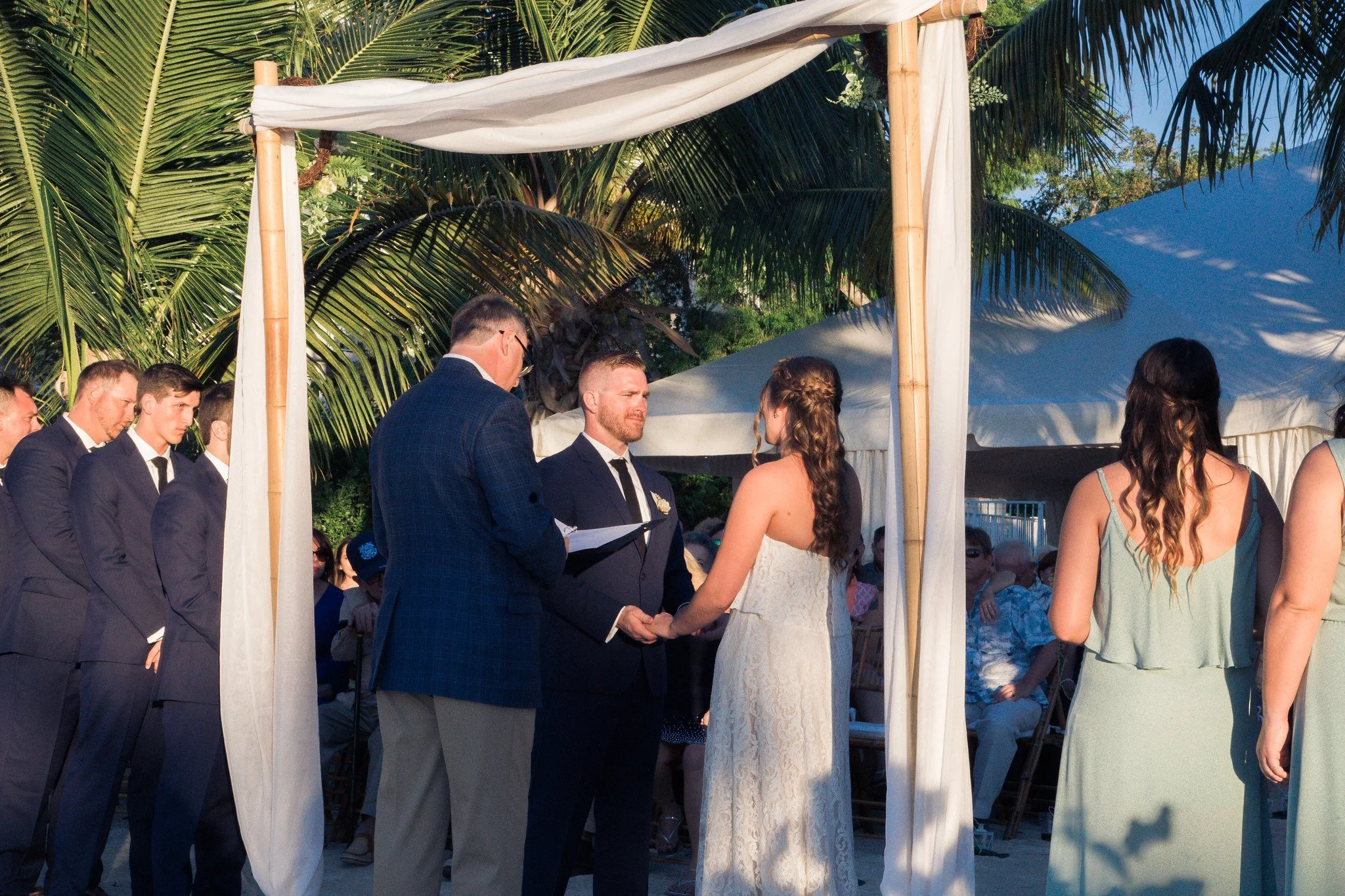 A couple getting married during an outdoor ceremony under a white canopy, with guests and palm trees in the background.