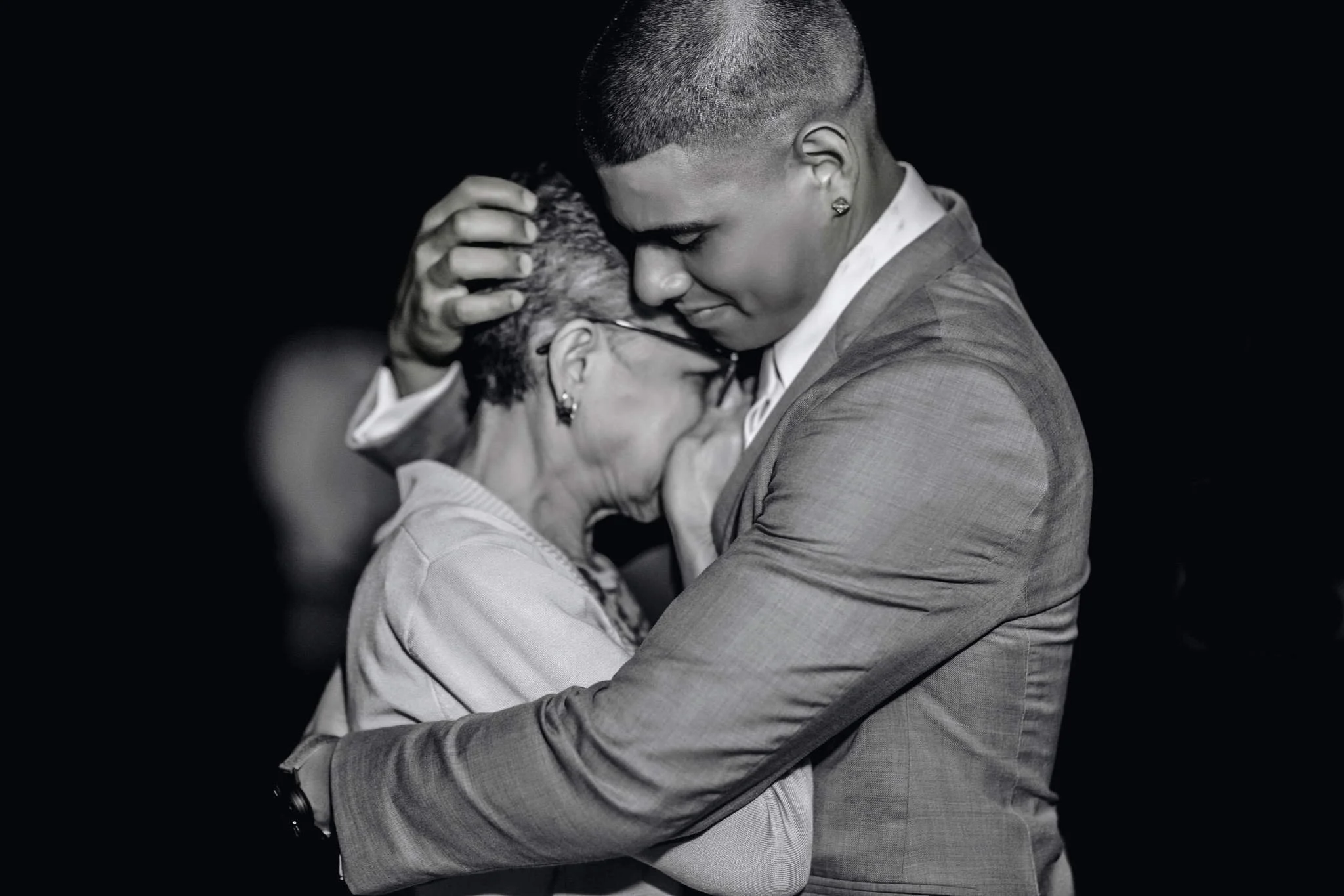 A young man and an older woman embrace tenderly, with their foreheads touching and eyes closed, in a heartfelt hug.