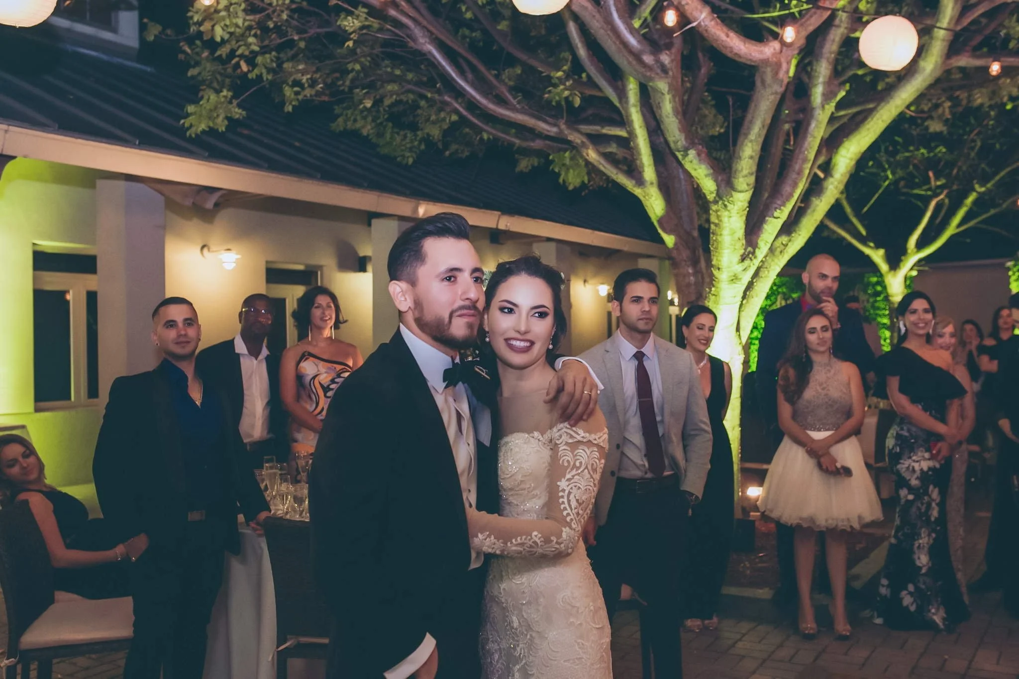 A bride and groom at a wedding reception, surrounded by friends and family, outdoors at night with trees and string lights.
