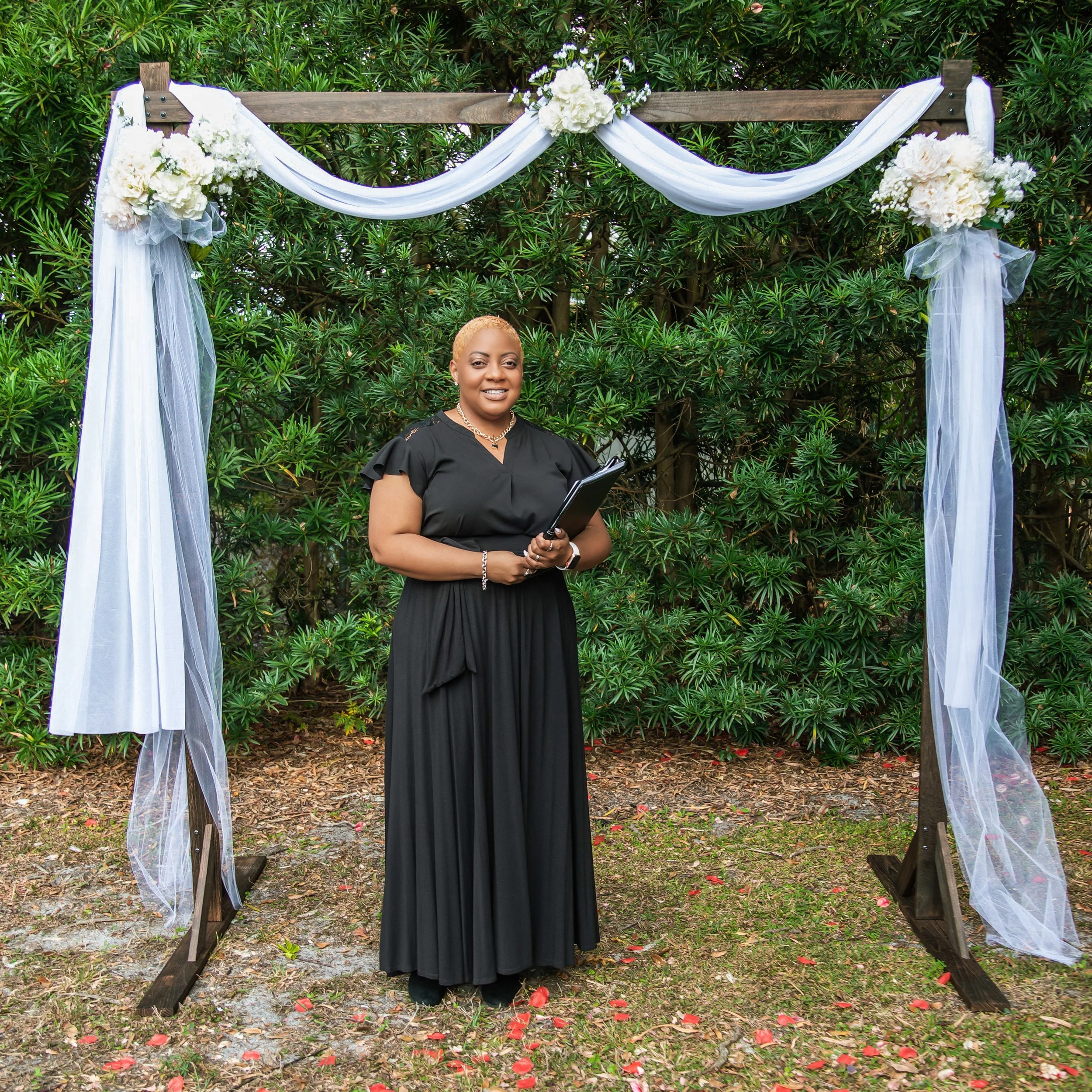 A woman in a black dress standing outdoors with a clipboard in front of a decorated wooden archway covered with white flowers and draped fabric, set against lush green bushes.