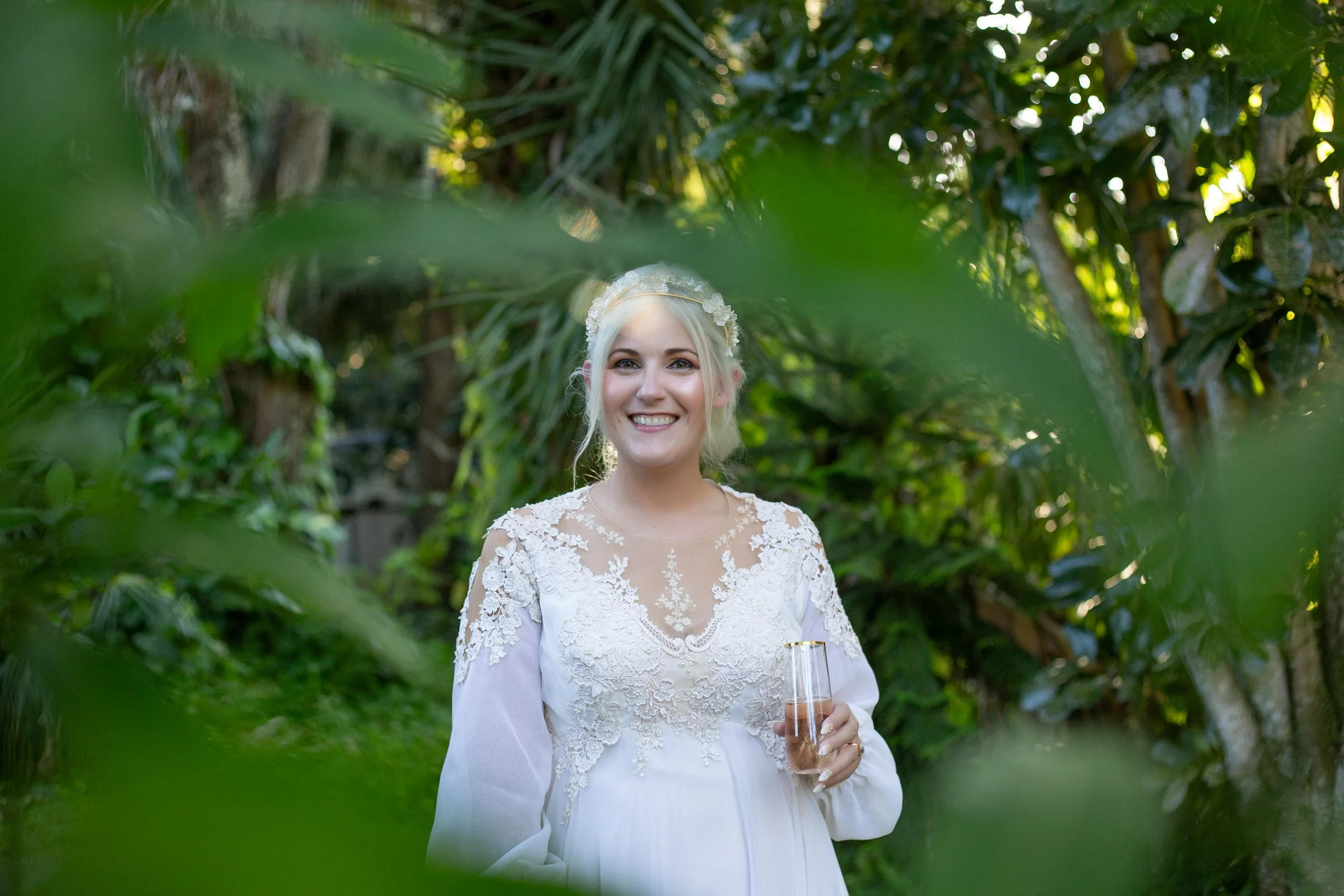 A smiling woman in a white lace dress holding a champagne flute, standing outdoors surrounded by green foliage.