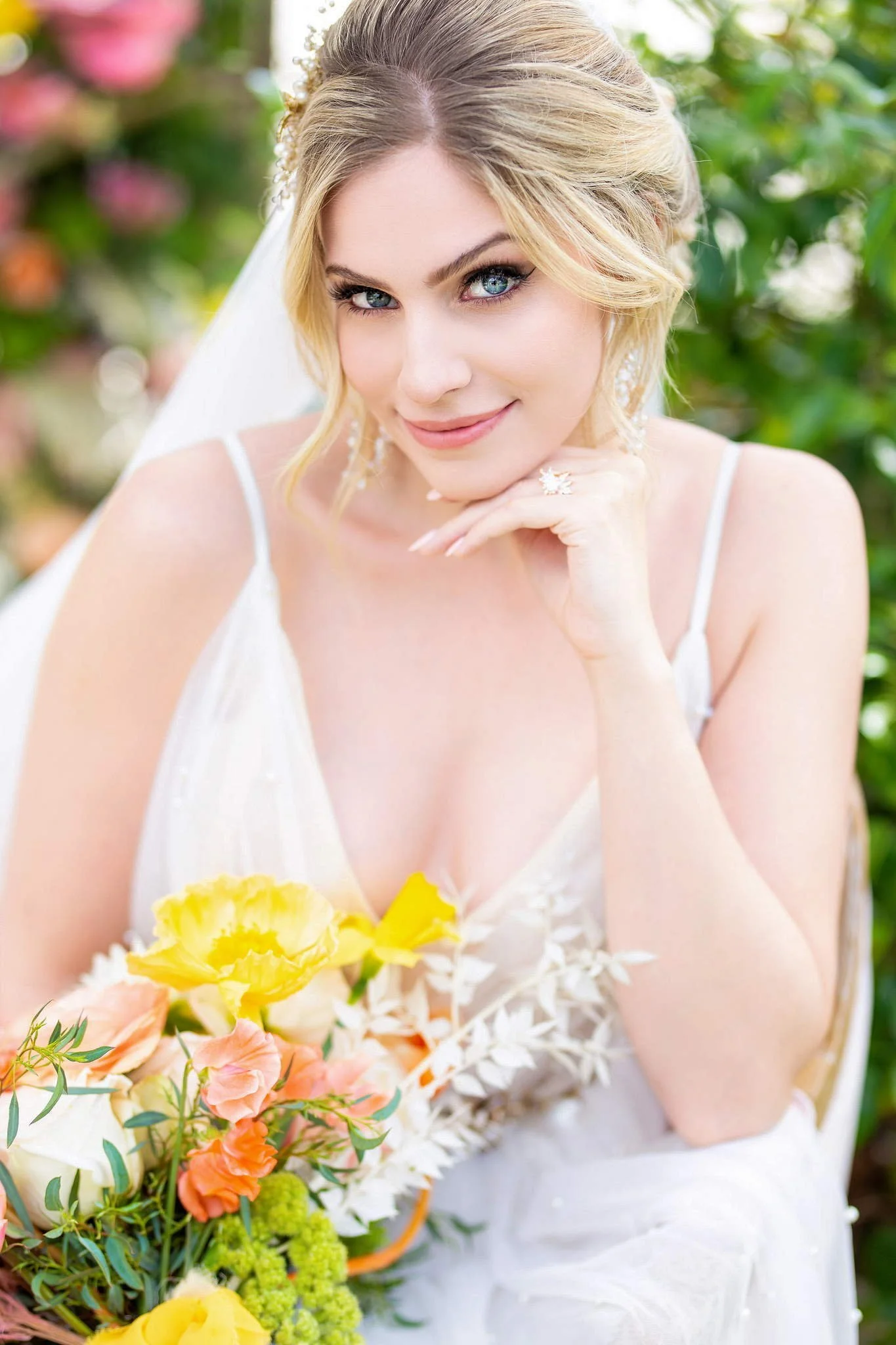 A smiling bride with blonde hair, wearing a white wedding dress with spaghetti straps, holding a colorful bouquet of yellow, peach, and pink flowers, standing outdoors with greenery background.