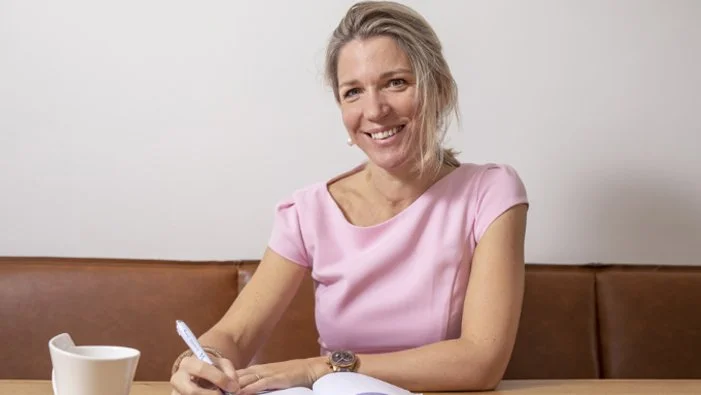 Vrouw in roze shirt schrijft in een notitieboekje aan tafel, naast een witte mok.