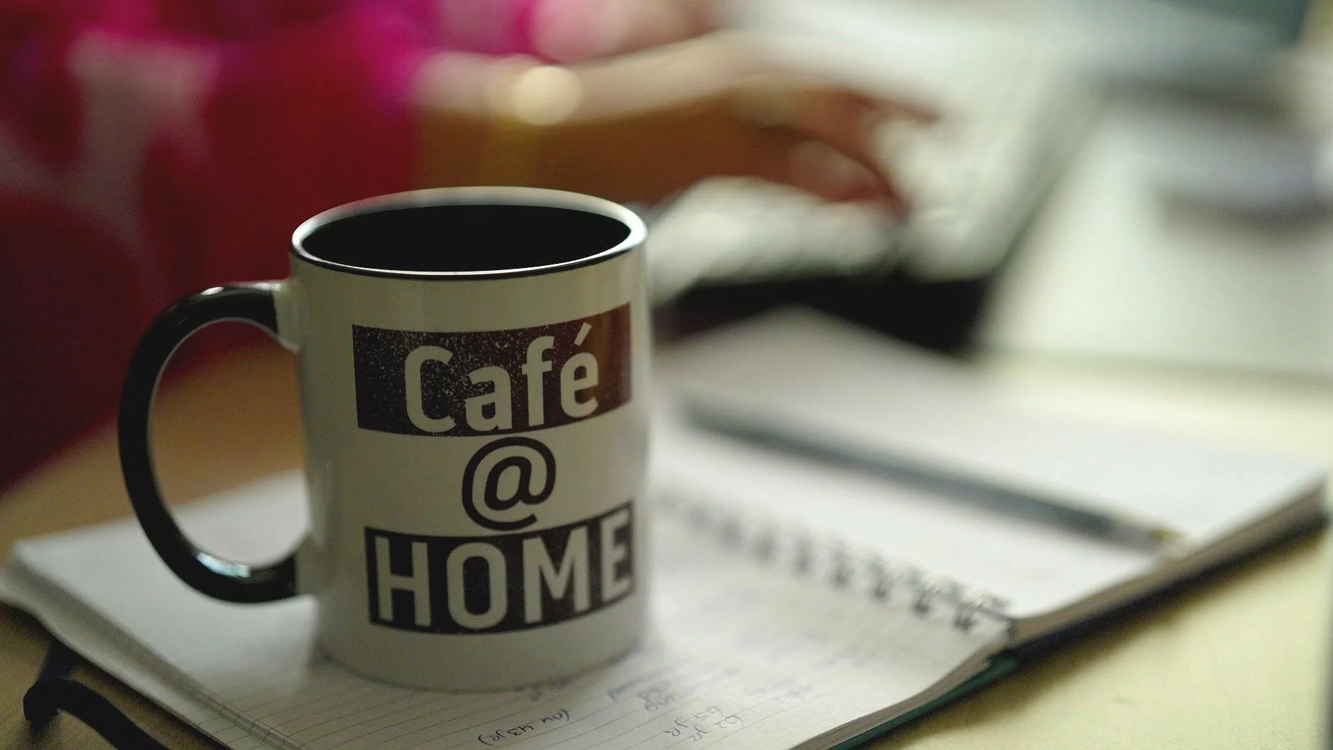 Koffiemok met tekst 'Café @ Home', notitieboek en pen op tafel, onscherpe achtergrond van persoon die typt op toetsenbord.